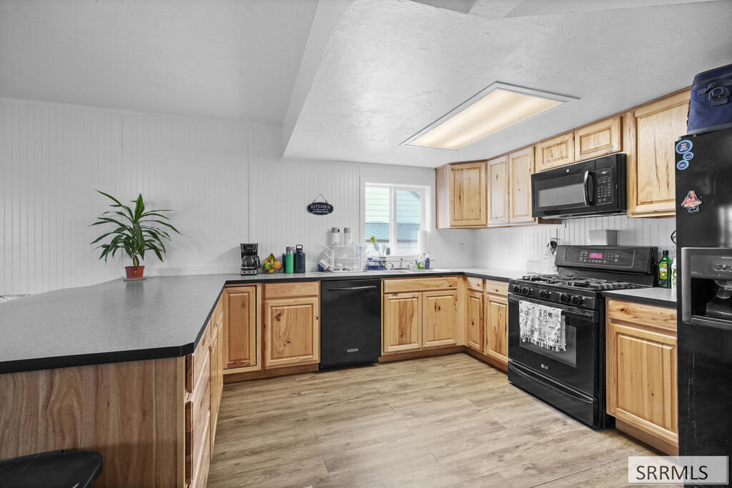 Kitchen with light wood cabinets, long gray counters, black appliances, and wood-look flooring.