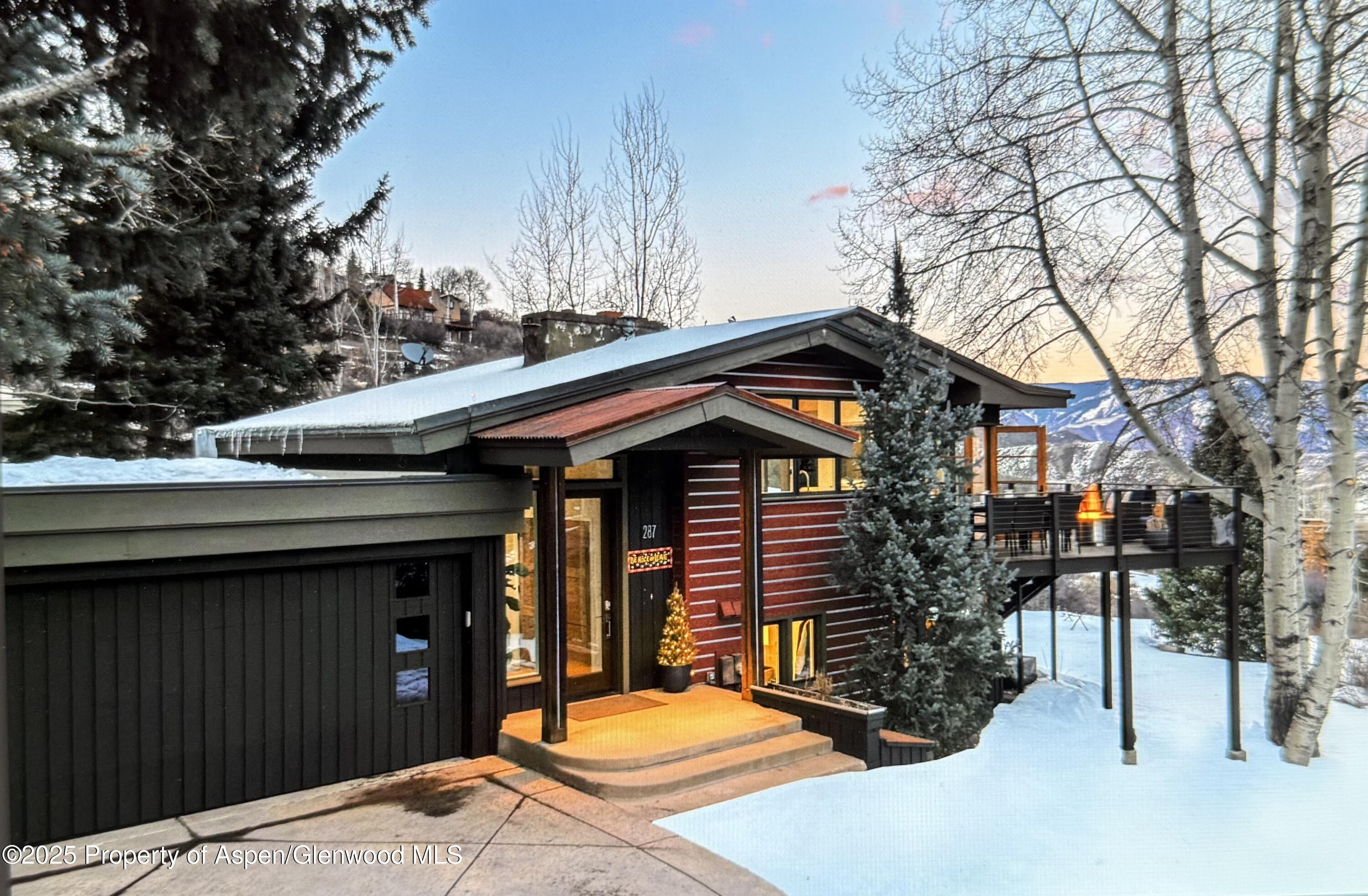 Contemporary red-sided home with an attached garage and elevated deck, surrounded by snow with mountains beyond.