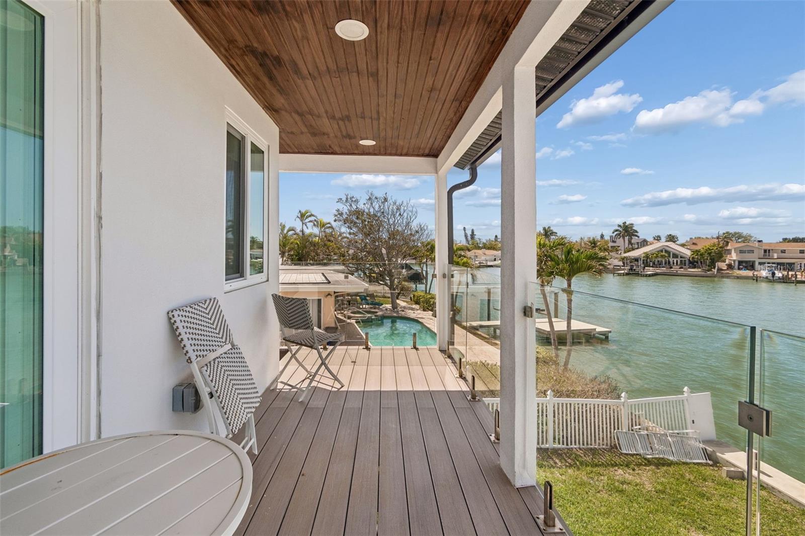 Covered balcony overlooking a canal, with glass railing, wood ceiling, and views of a pool and neighboring homes.