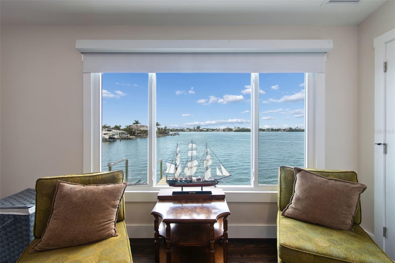 Living room with a large waterfront window, two green chairs, and a model sailing ship on the coffee table.