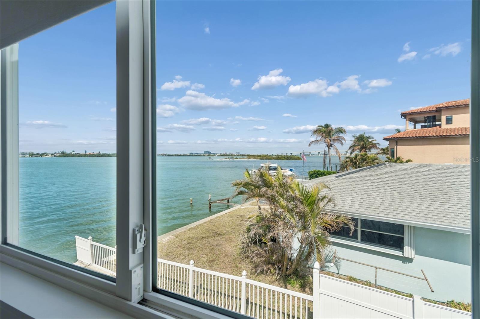 Waterfront view through a window, with boats, palm trees, and neighboring homes along the shore.