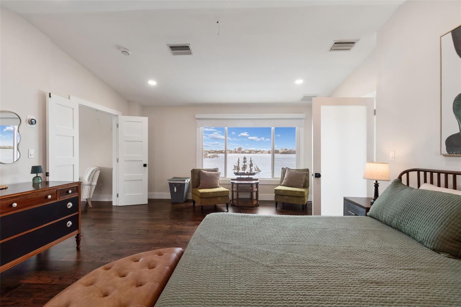 Furnished bedroom with a water view, dark wood floors, and a sitting area by the window.