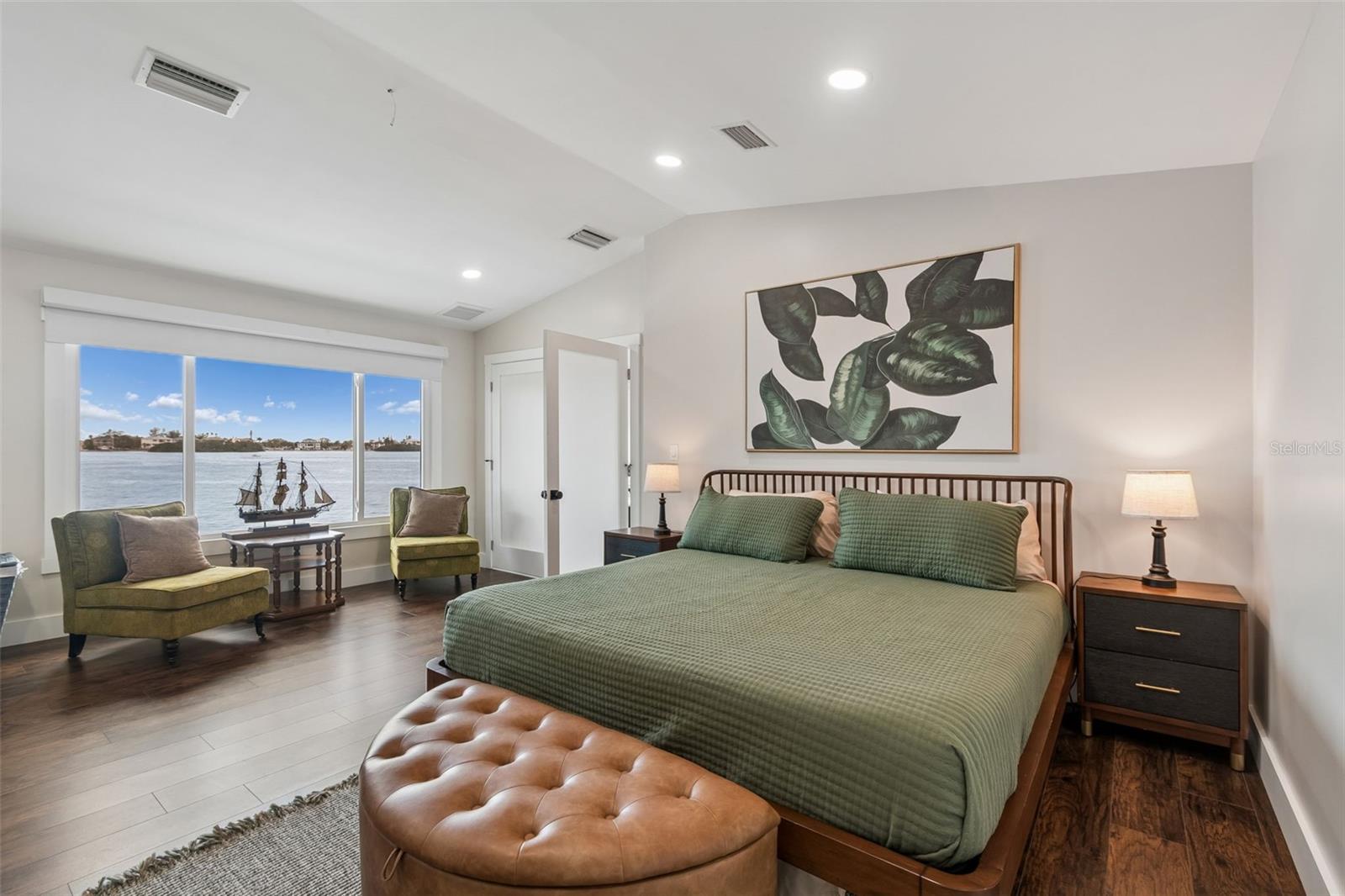 Bedroom with green bedding and seating beside large windows overlooking the water.