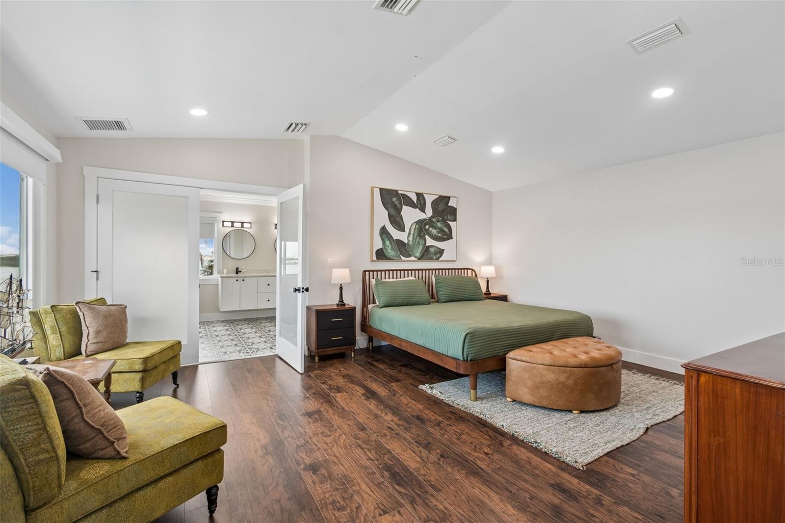 Furnished bedroom with a green bed, seating area, dark wood floors, and open doors to an en-suite bathroom.