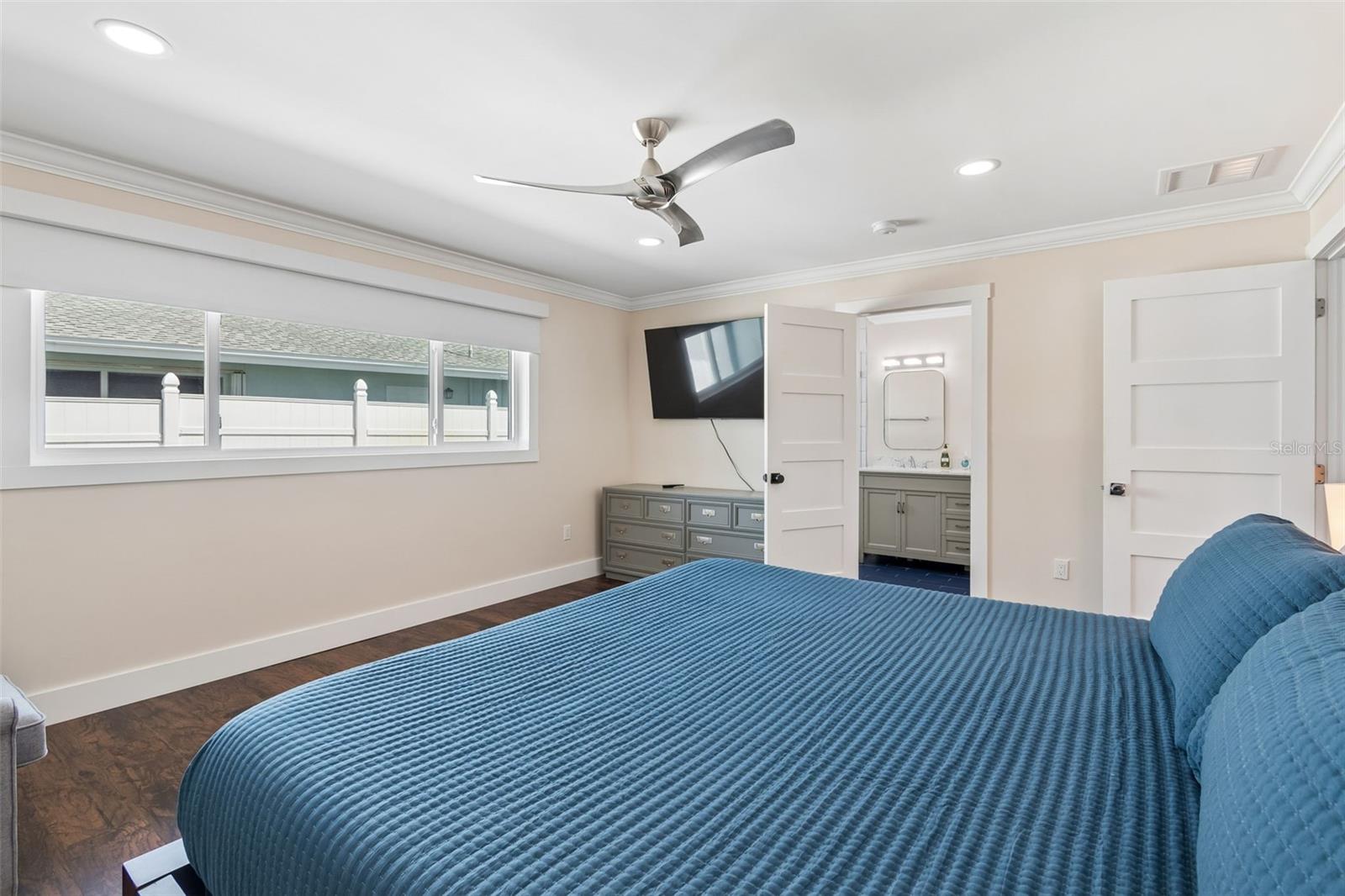 Bedroom with a blue quilted bed, wide horizontal window, ceiling fan, and open doorway to a bathroom.