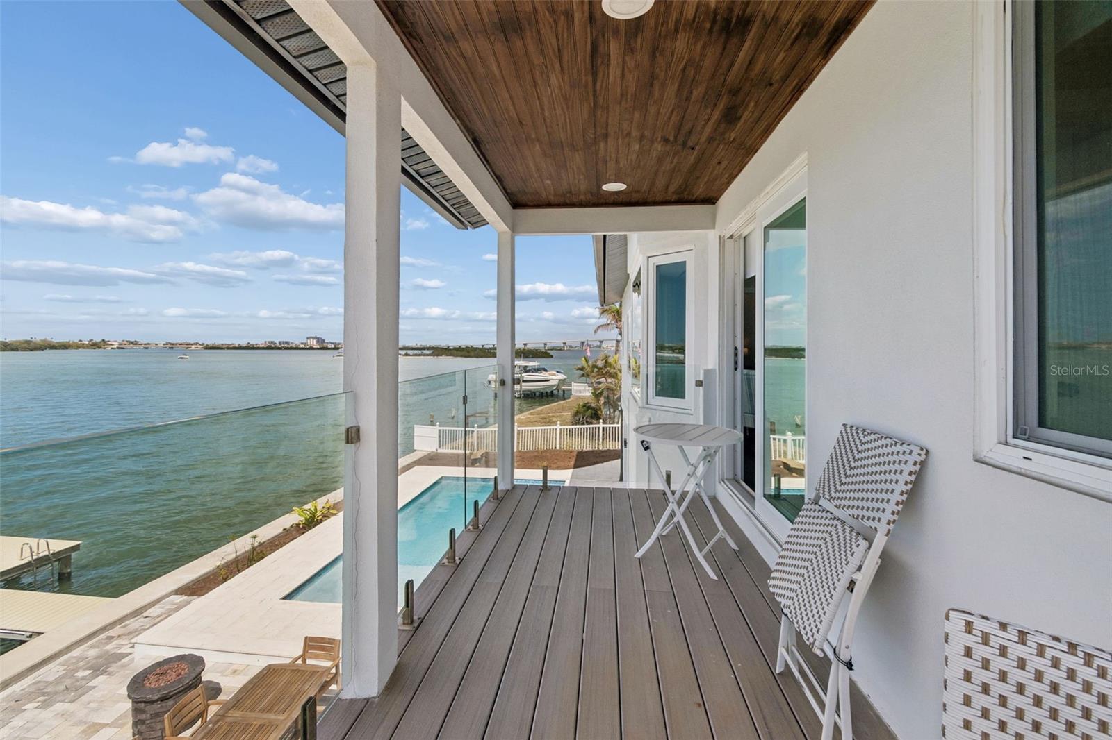 Covered waterfront balcony with glass railing, wood ceiling, and views across the water.