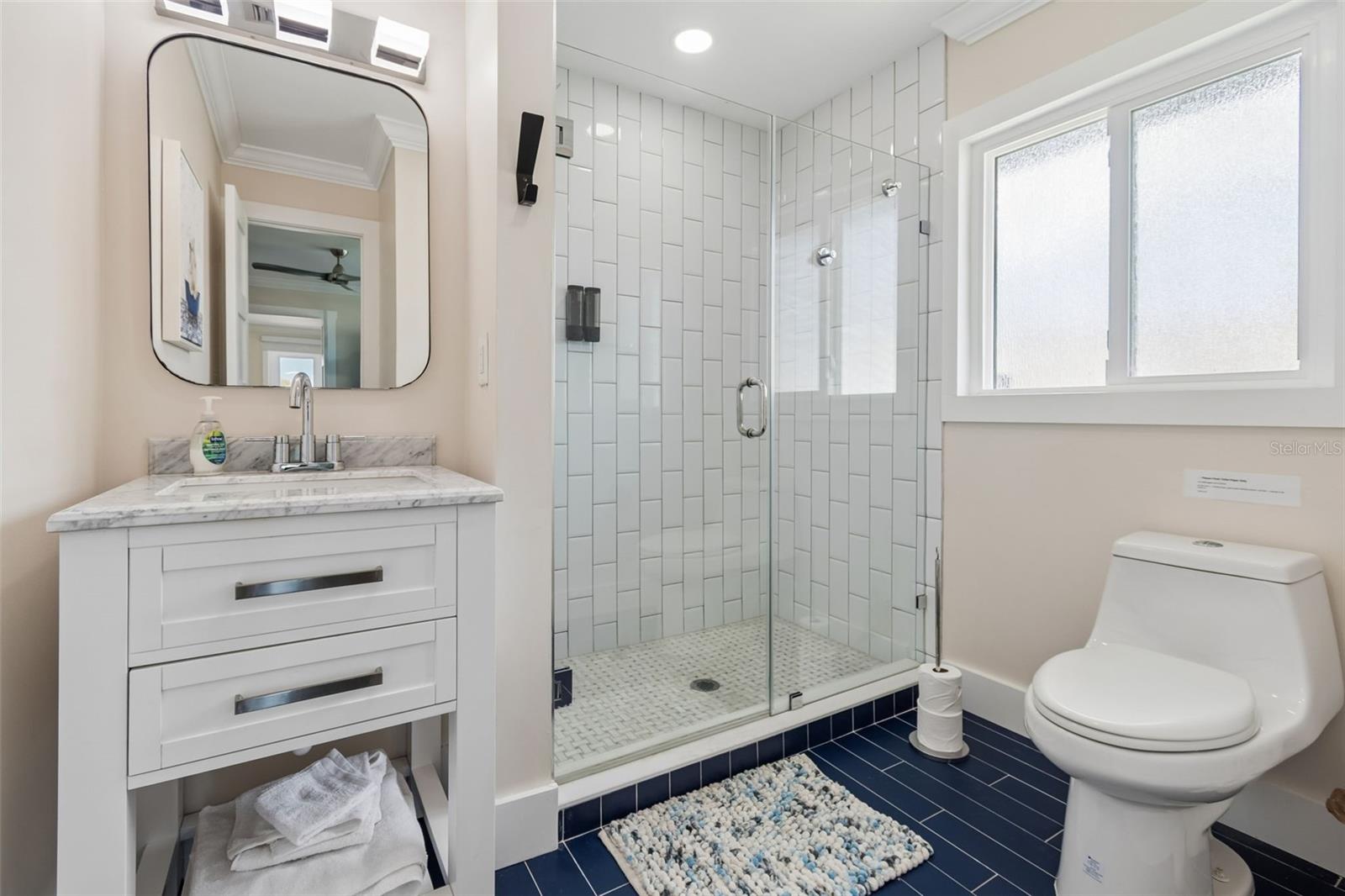 Bathroom with a glass-enclosed tiled shower, white vanity, frosted window, and navy floor tile.