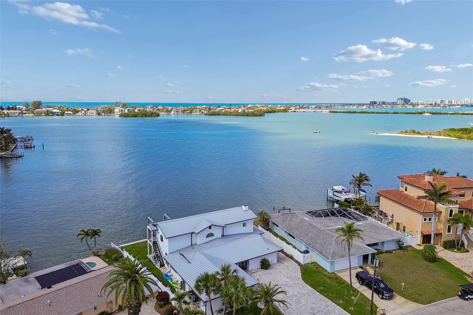 Aerial view of waterfront homes along a broad bay, with docks and distant coastal buildings.