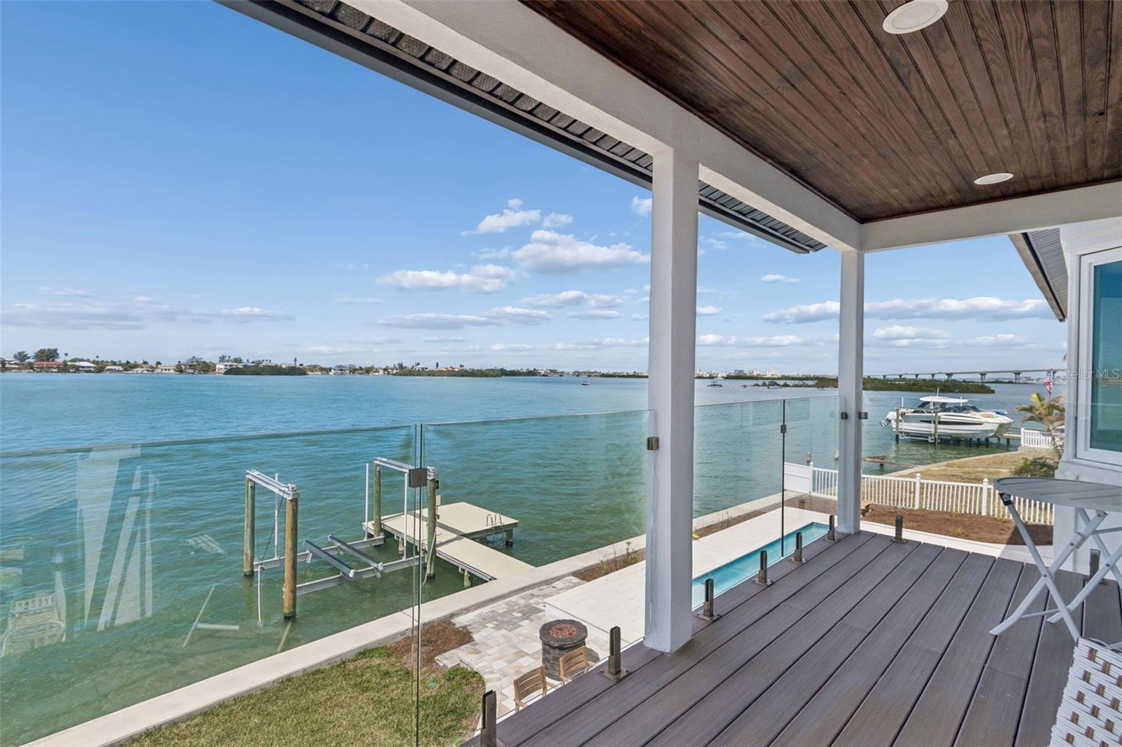 Covered waterfront deck overlooking the bay, with glass railing and boats at nearby docks.