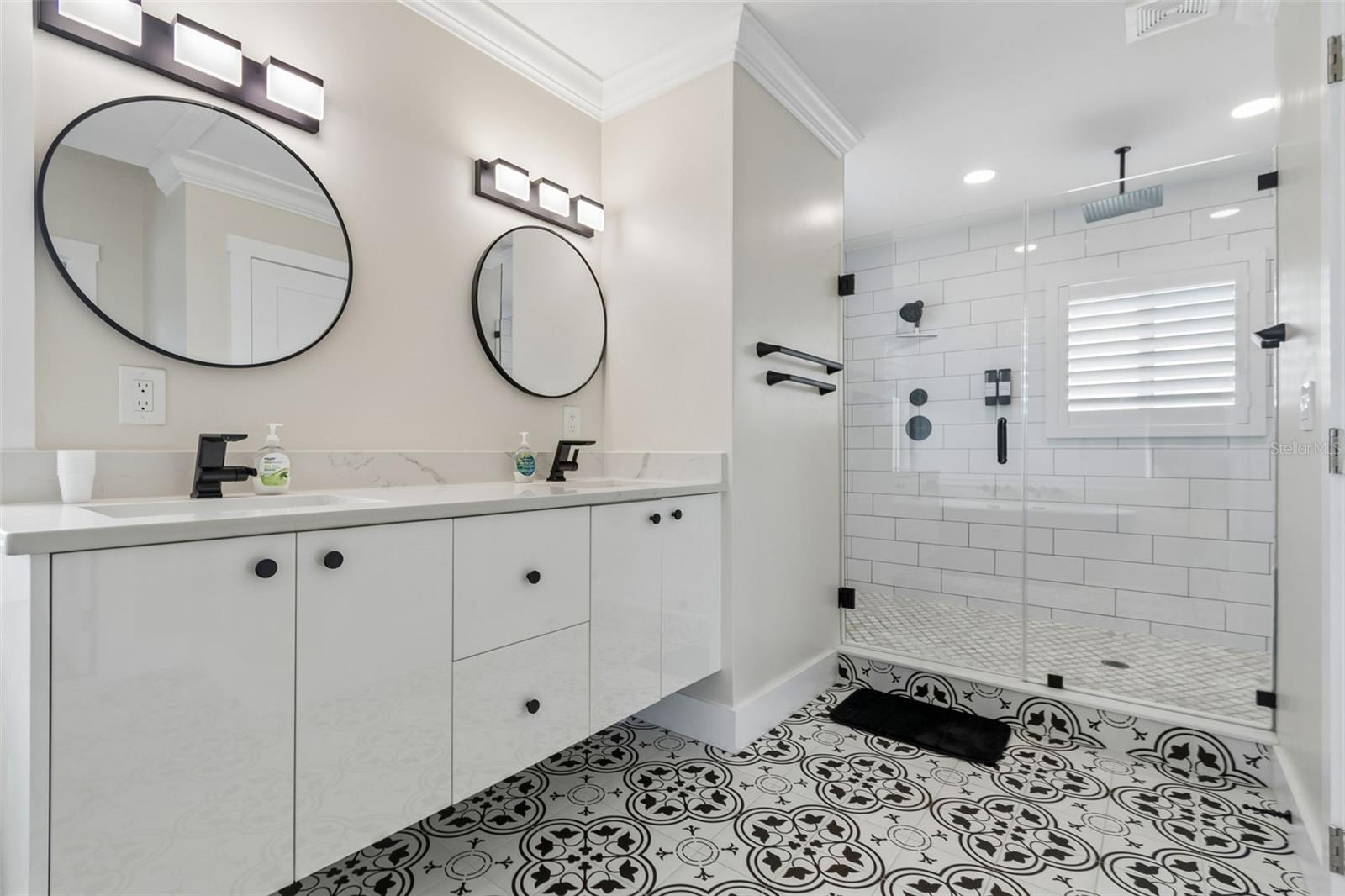 Bright bathroom with a glass walk-in shower, double mirrors, and a bold patterned tile floor.