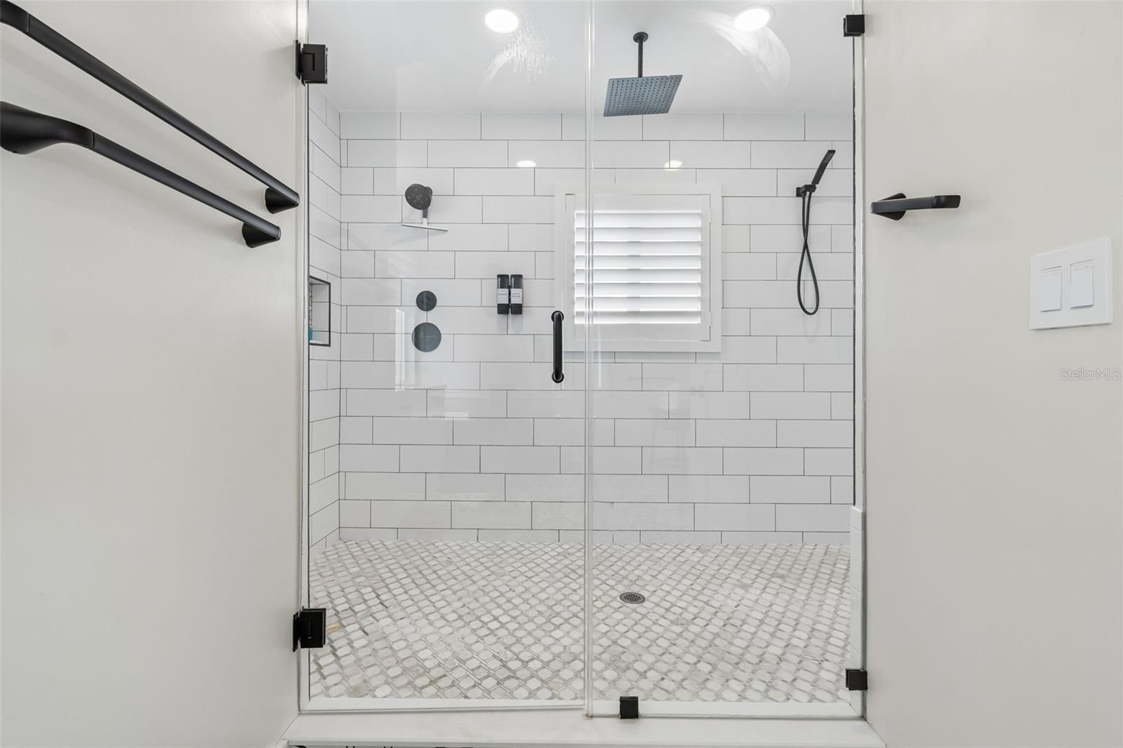 Walk-in shower with white subway tile, a mosaic floor, black fixtures, and a shuttered window.