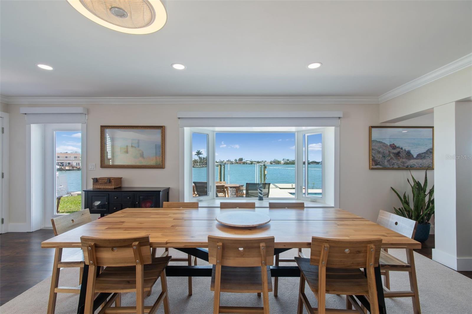 Waterfront dining room with a long wooden table facing wide windows and a dock across the bay.