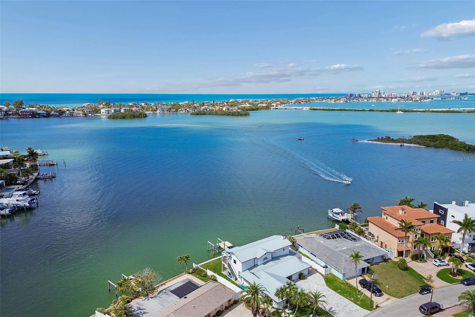 Aerial view of waterfront homes along a broad bay, with a boat crossing the water and a distant skyline.