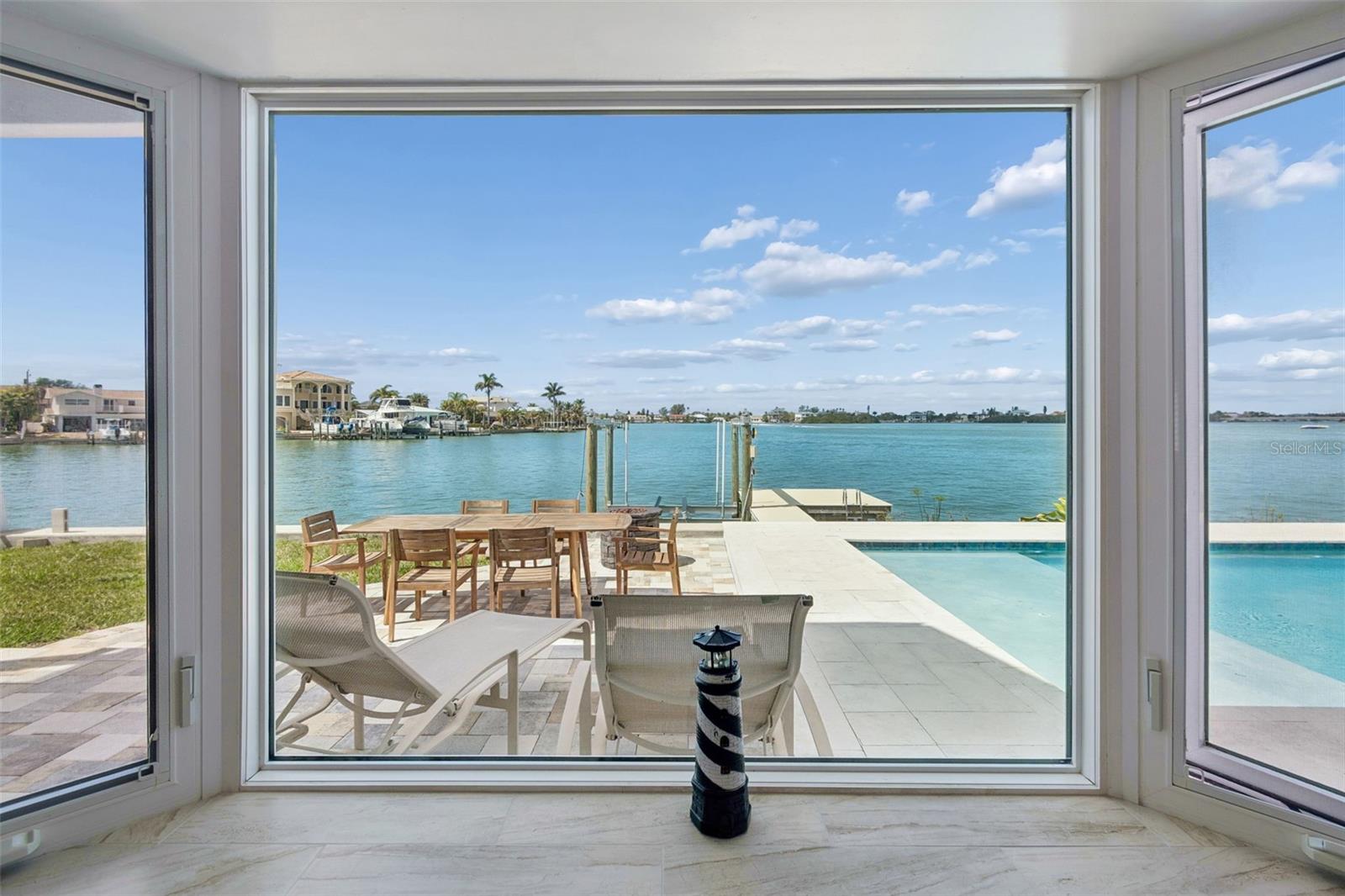 Waterfront patio and pool viewed through large windows, with a dock and homes across the water.