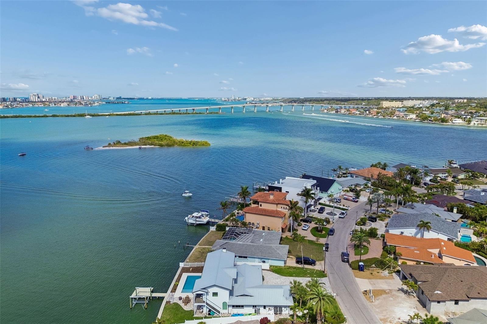 Aerial view of waterfront homes and docks, with a bridge, small island, and distant city skyline.