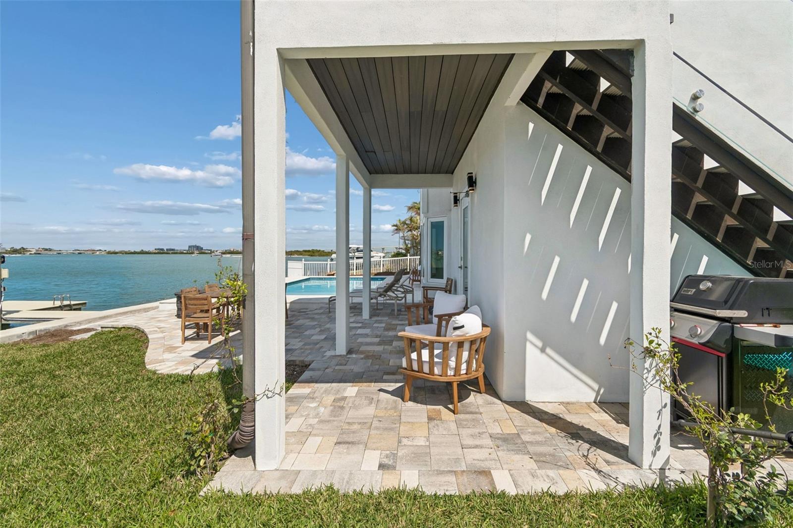 Waterfront patio with stone pavers, wooden seating, a grill, and a pool beside the water.