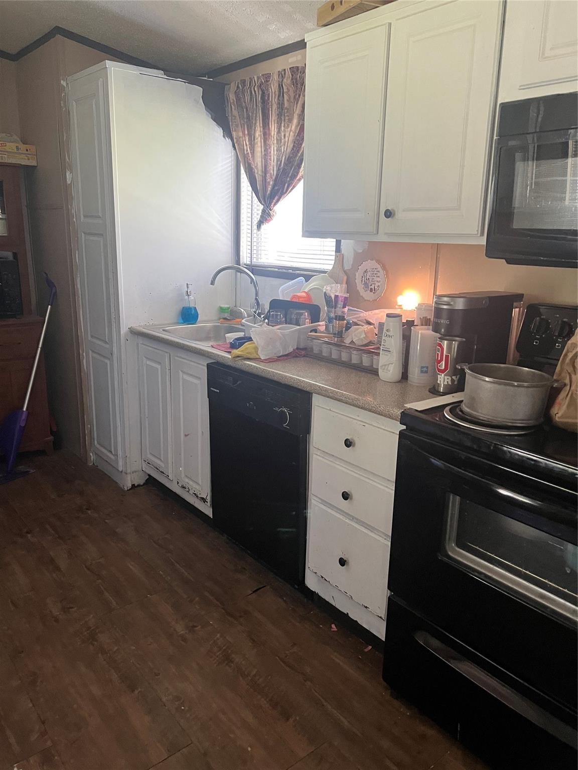 Kitchen with white cabinets, a sink beneath a curtained window, and black appliances.