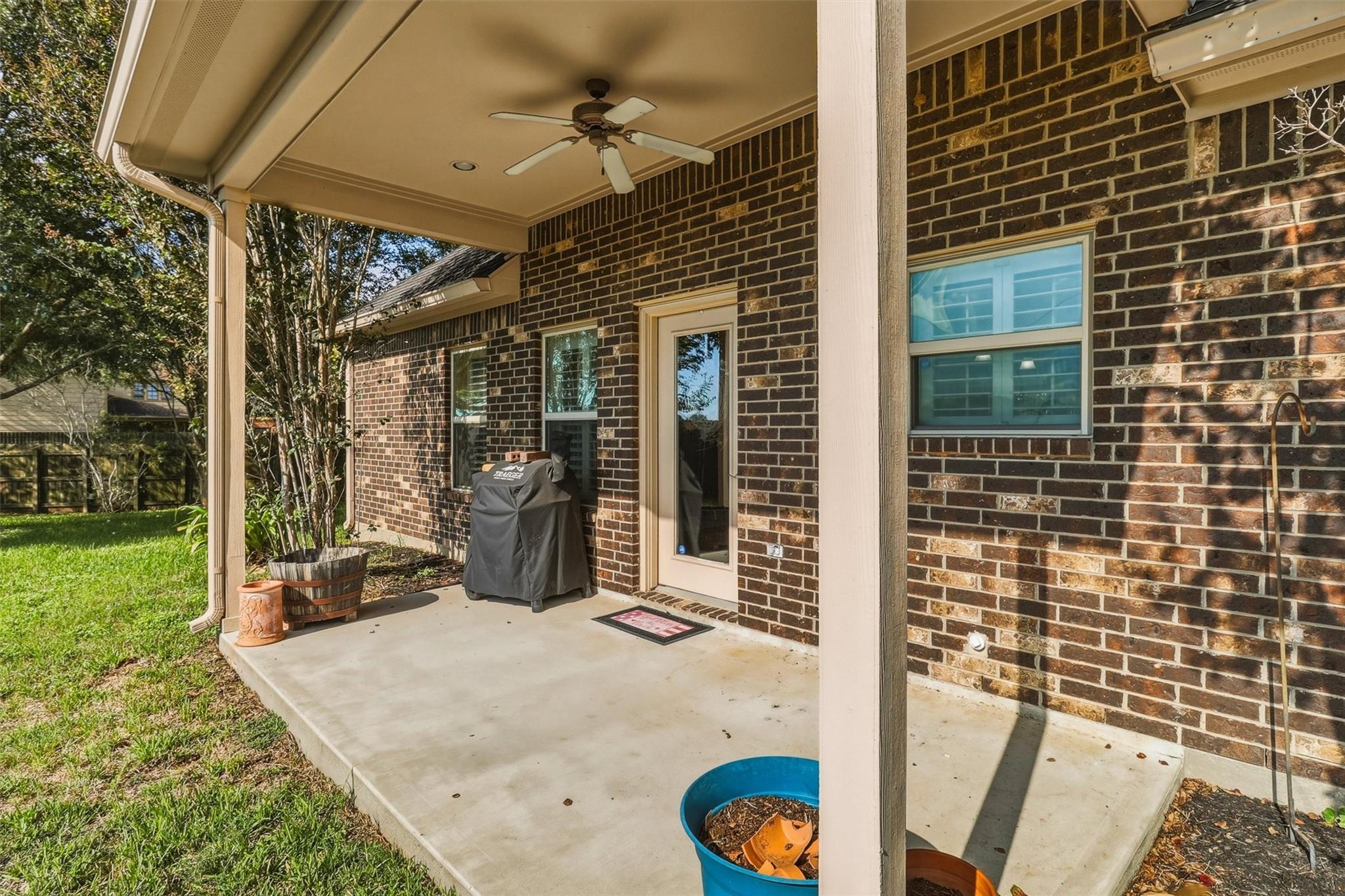 Covered brick-home patio with a ceiling fan, glass door, and grill.
