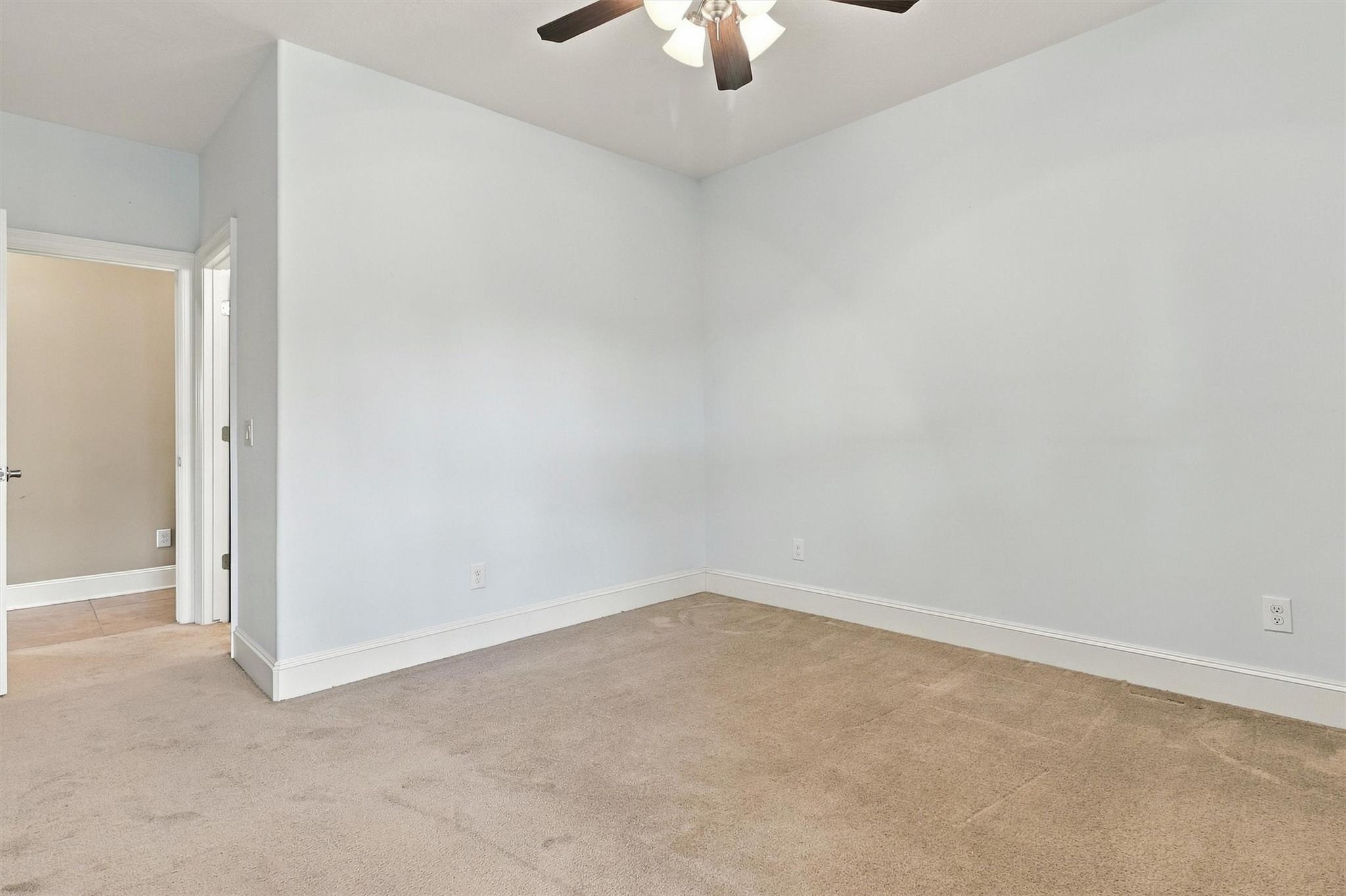 Empty carpeted room with pale walls, a ceiling fan, and an open doorway to a tiled hall.