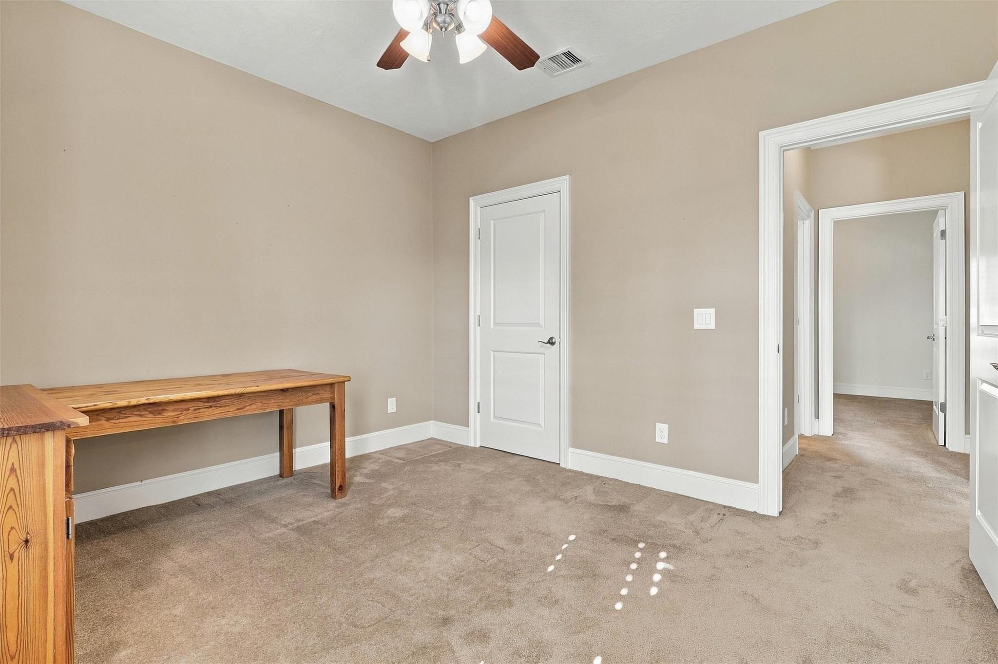 Interior room with a wooden table, beige carpet, ceiling fan, and doorway to a hallway.