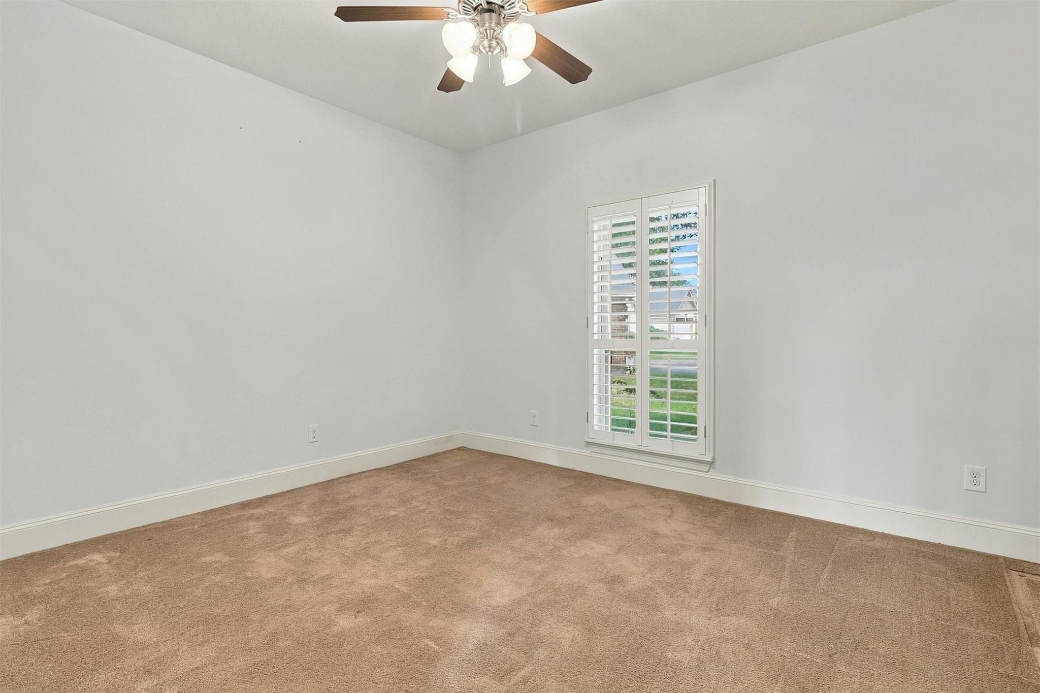 Empty bedroom with beige carpet, a ceiling fan, and a shuttered window.