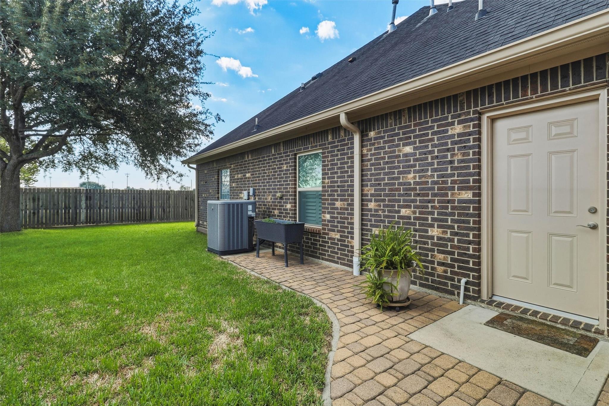 Brick house side entrance with a white door, curved paver walkway, and fenced lawn beneath a large tree.