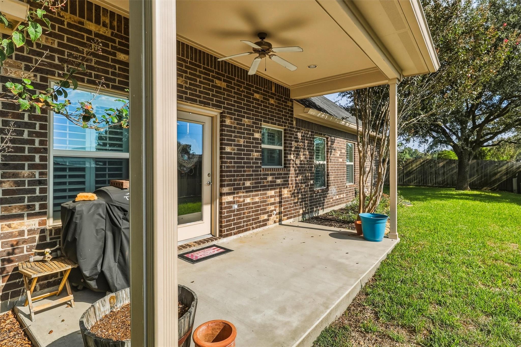 Covered brick patio with a ceiling fan, grill, and view of the fenced backyard.