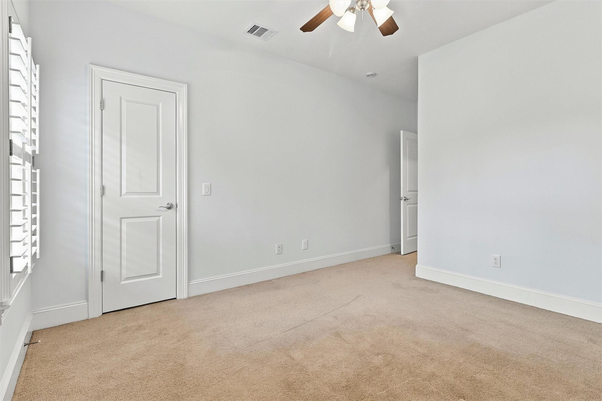 Empty bedroom with beige carpet, plantation shutters, and a ceiling fan.