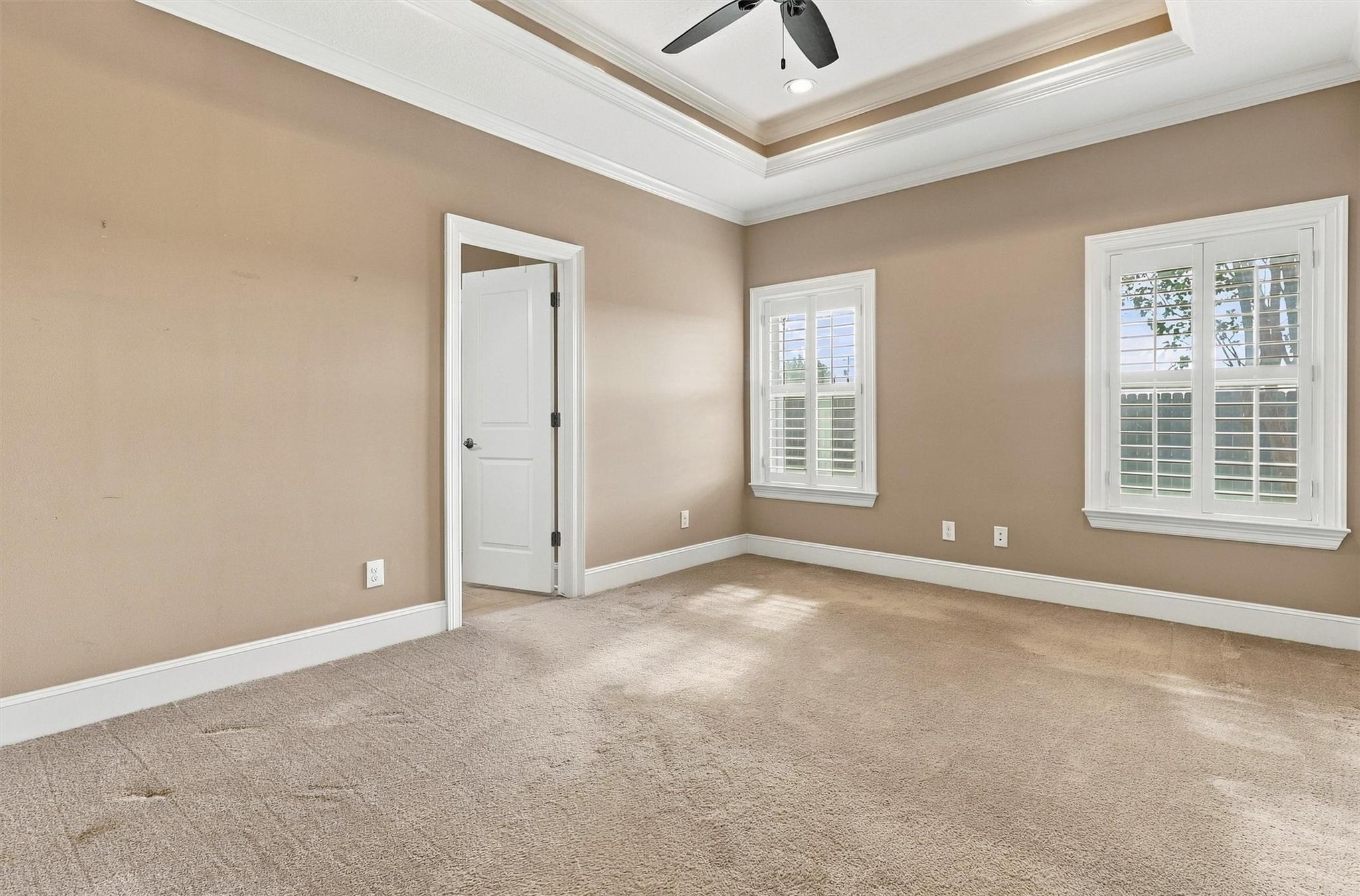 Empty carpeted room with a tray ceiling, ceiling fan, and two shuttered windows.