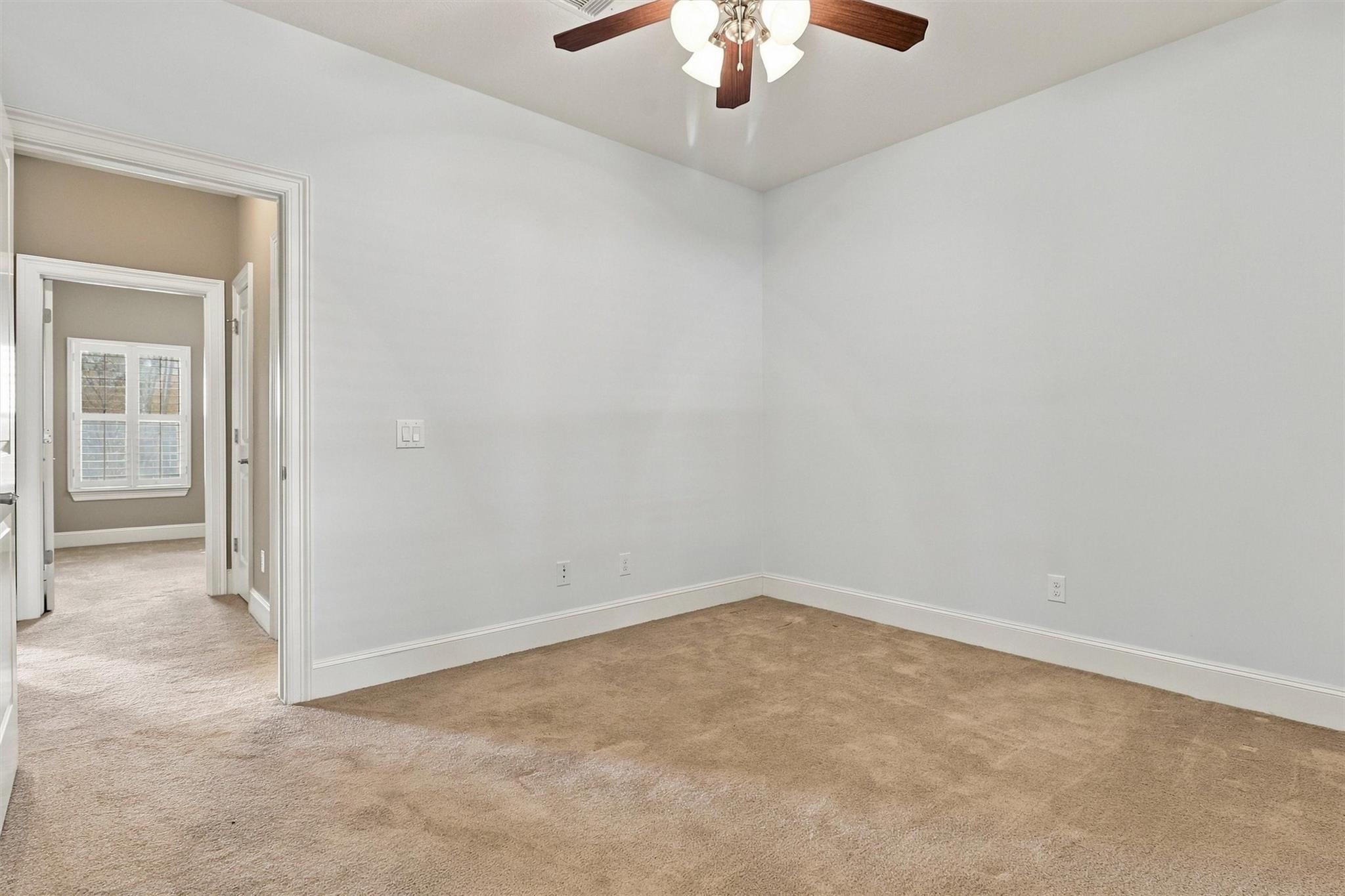 Vacant carpeted bedroom with a ceiling fan and an open doorway to a room with a window.
