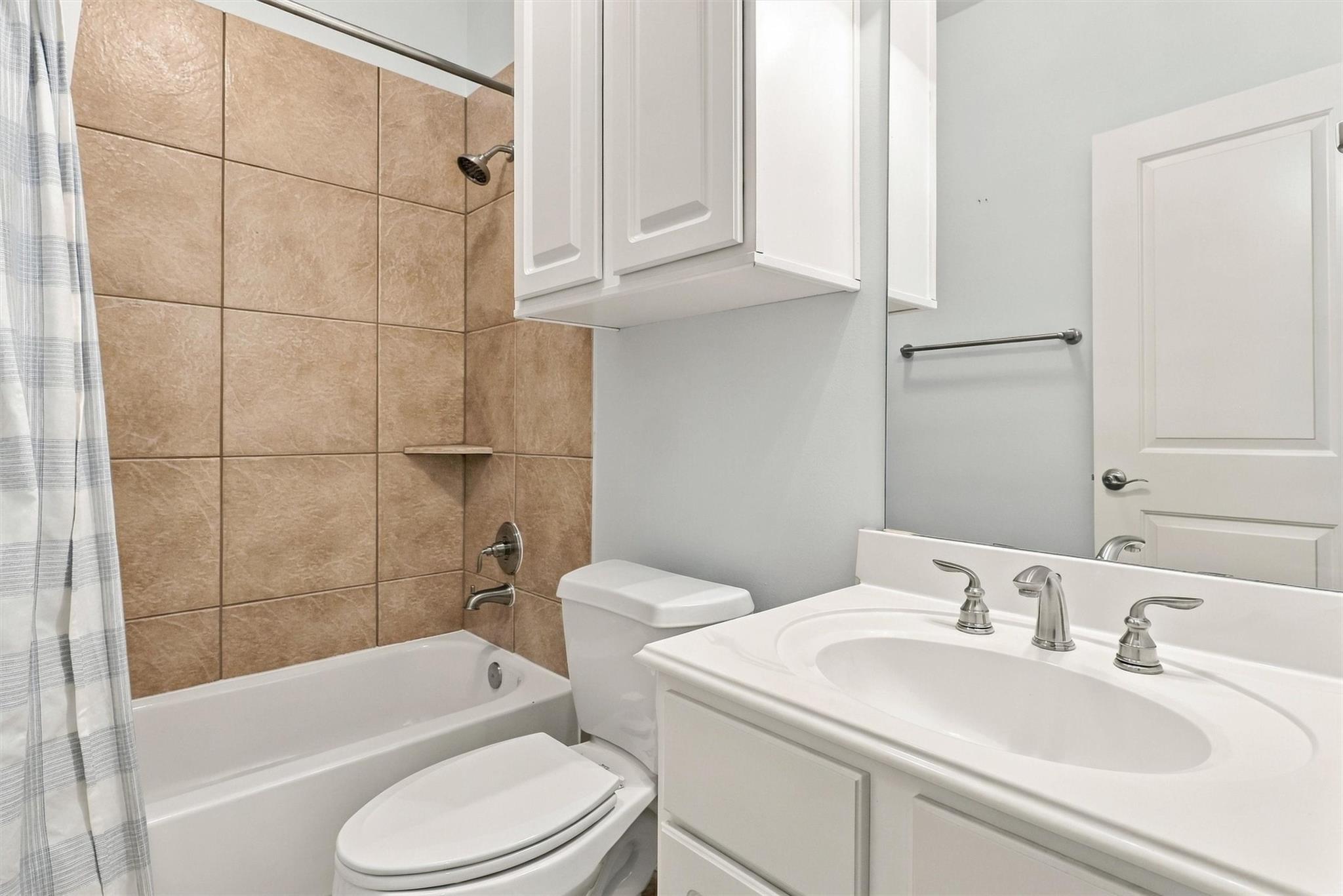 Bathroom with a tan tiled tub and shower, white toilet, and vanity sink.