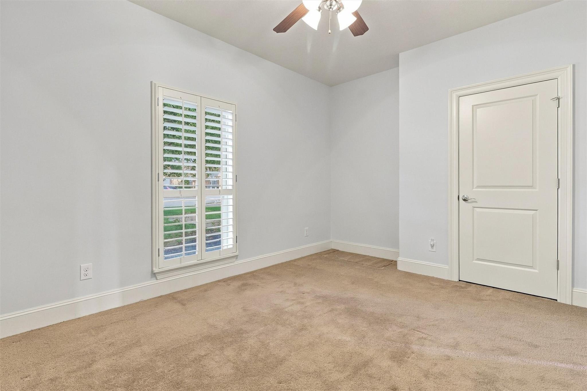 Empty carpeted room with plantation shutters, a white paneled door, and a ceiling fan.