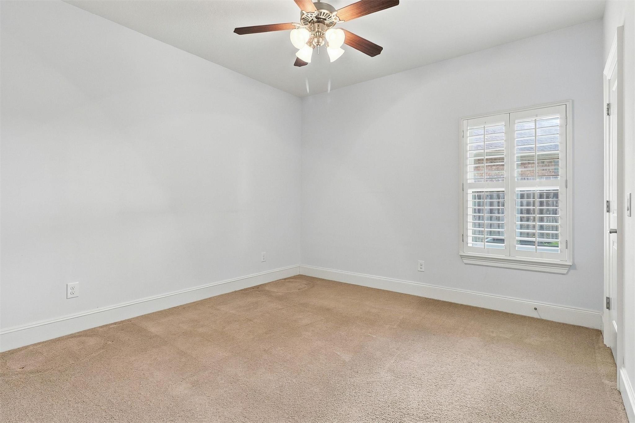 Empty carpeted room with a ceiling fan and a shuttered window.
