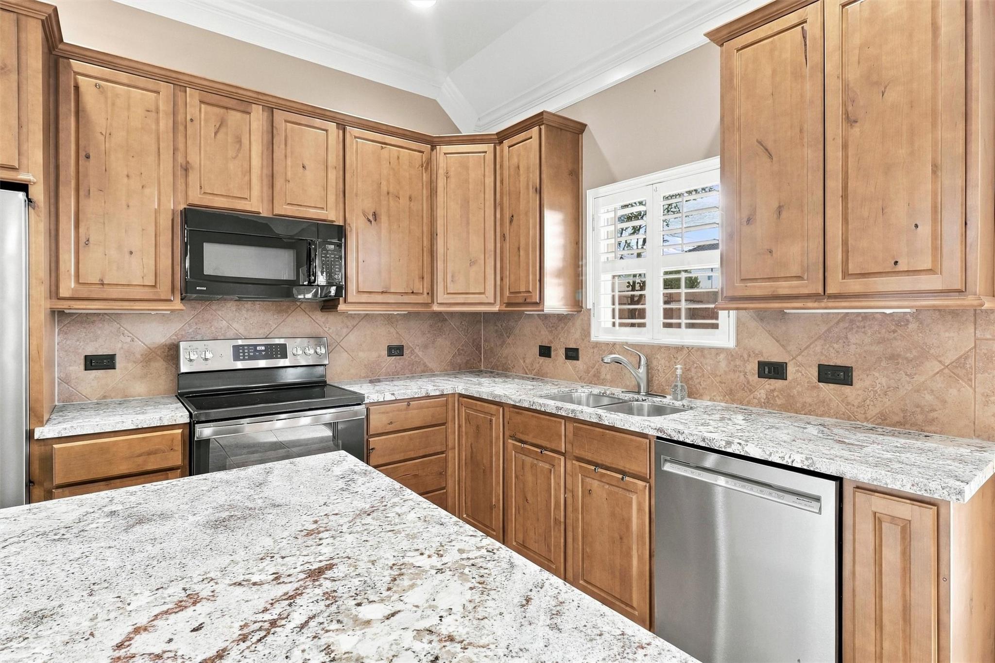Kitchen with warm wood cabinets, granite counters, stainless steel appliances, and a sink beneath a shuttered window.