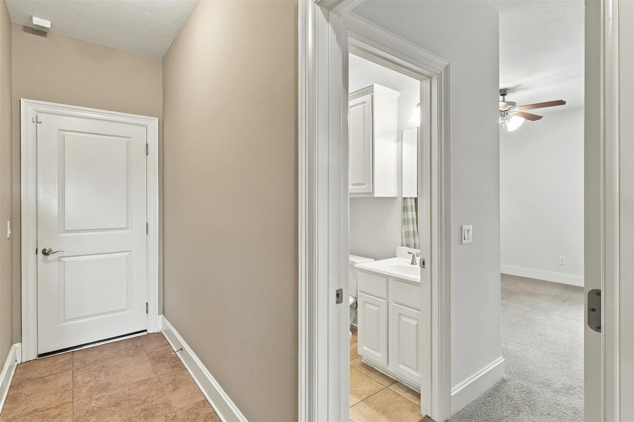 Hallway with tile floor, bathroom vanity, and adjoining carpeted room with ceiling fan.