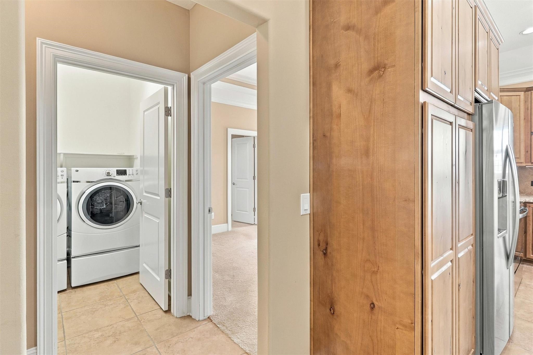 Laundry closet with stacked washer and dryer beside wood kitchen cabinets and a stainless steel refrigerator.