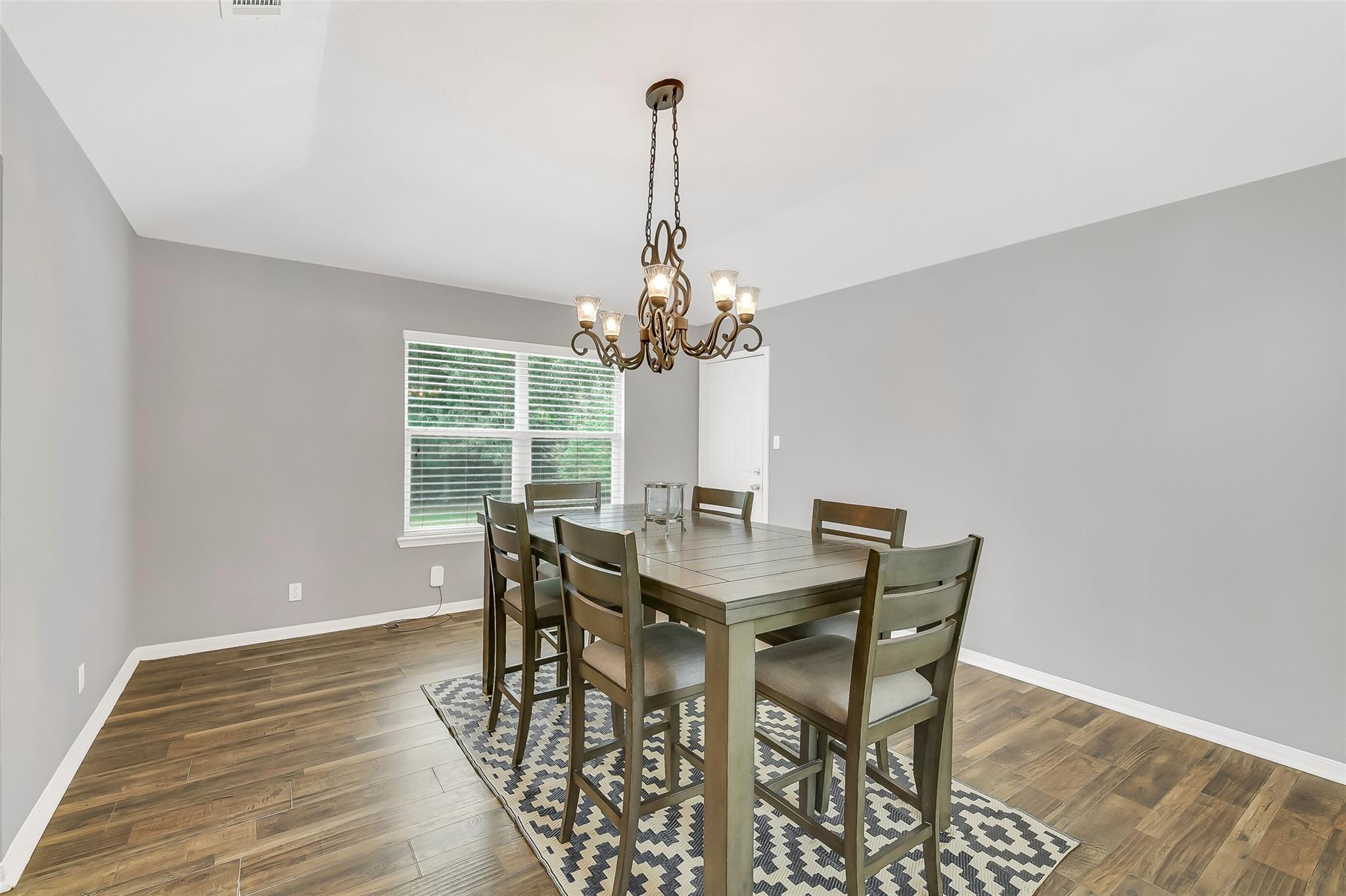Dining room with a large table, patterned rug, and chandelier beside a window.