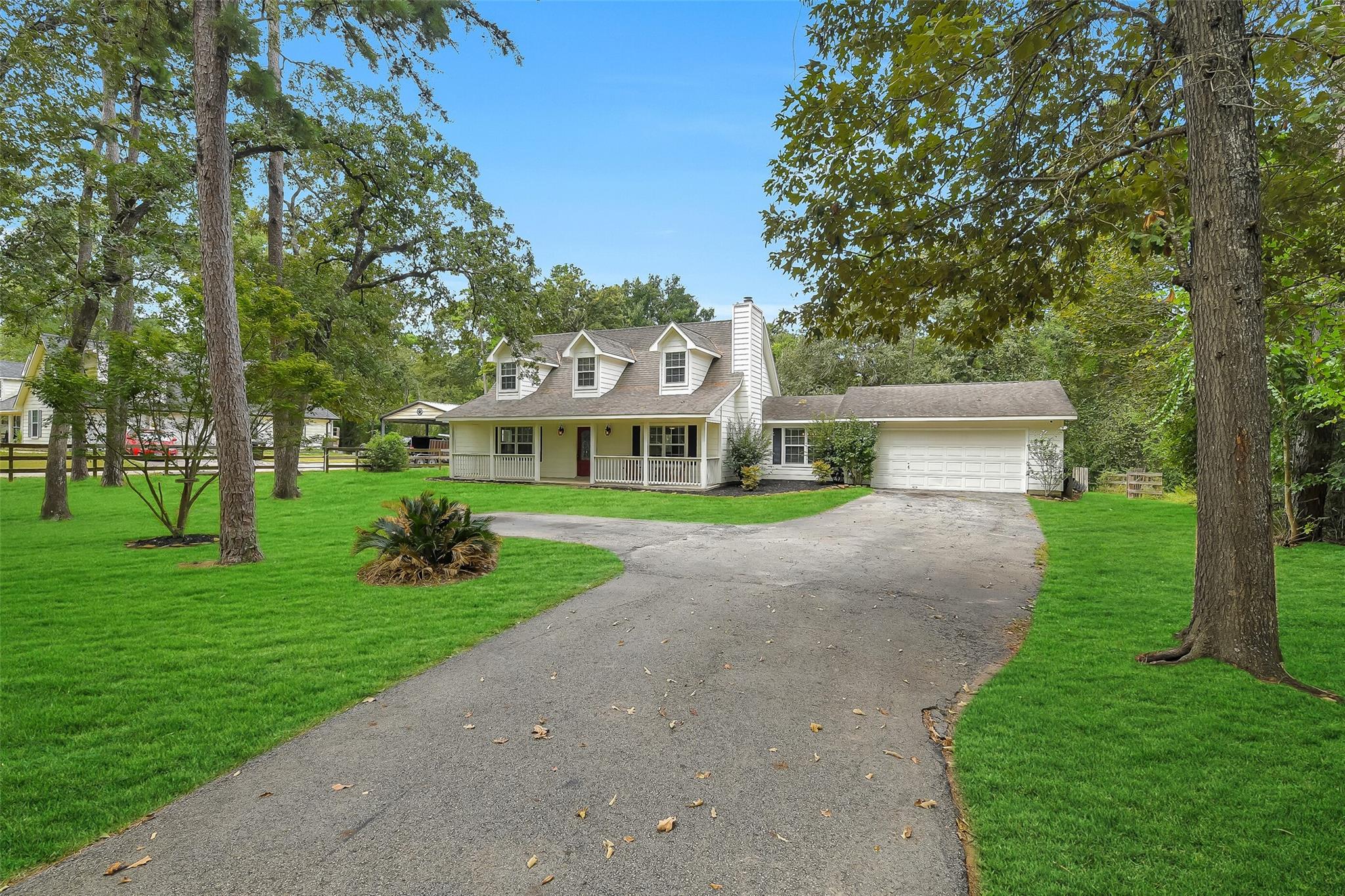 White two-story home with dormers, a covered porch, attached garage, and curved driveway beneath mature trees.