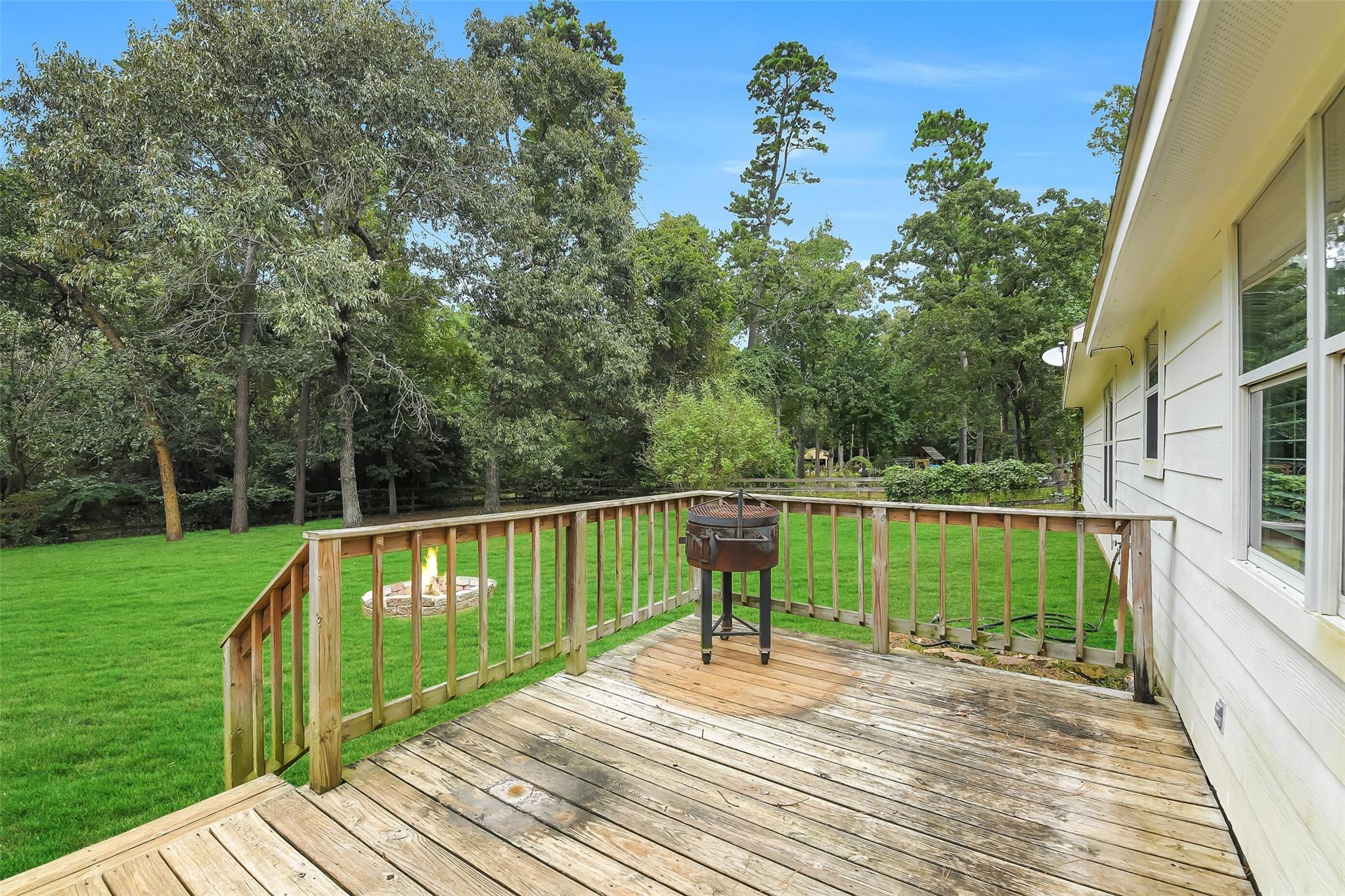Wooden backyard deck beside a white house, with a grill, lawn, mature trees, and a fire pit.