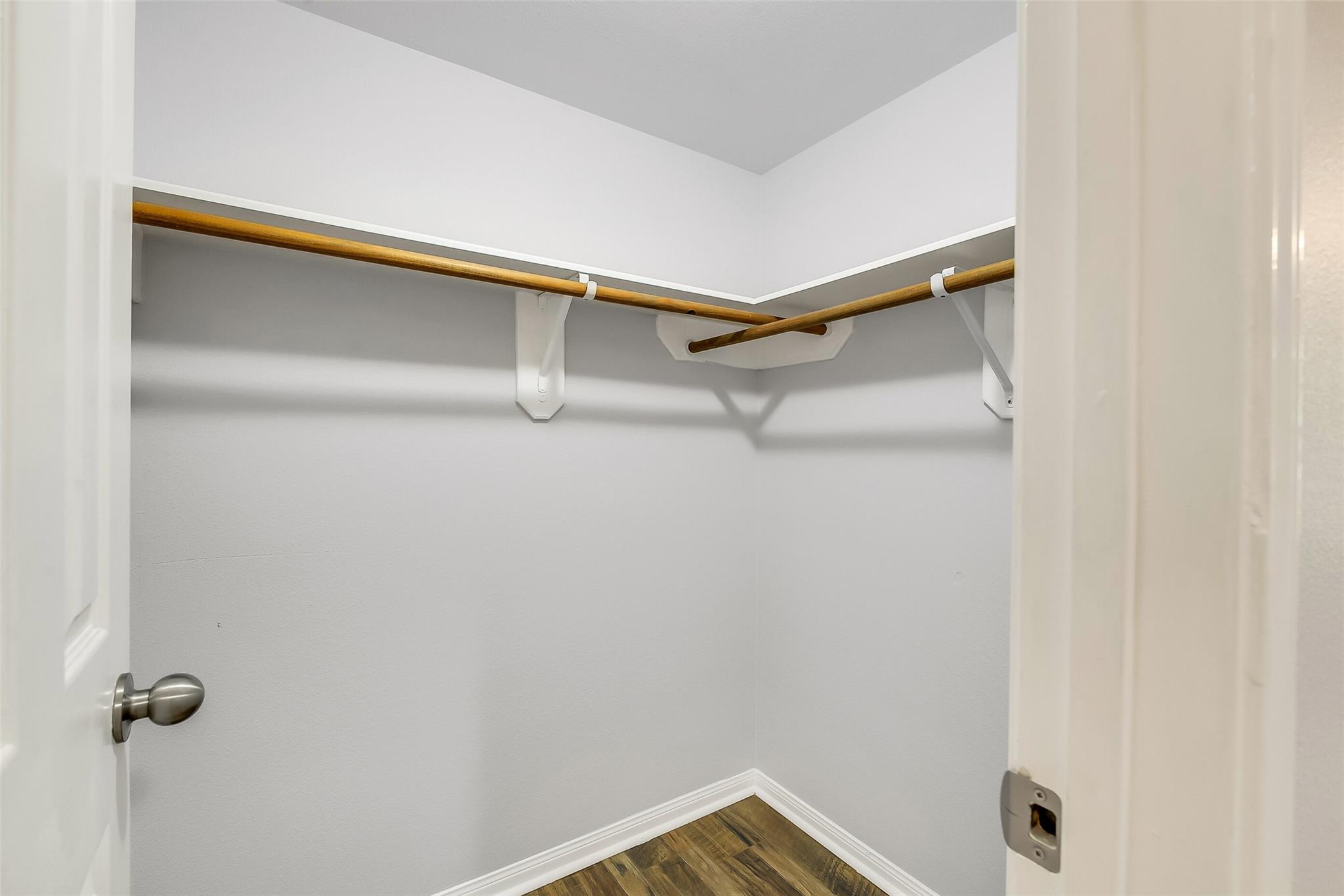 Empty closet with wooden hanging rods on two walls and wood-look flooring.