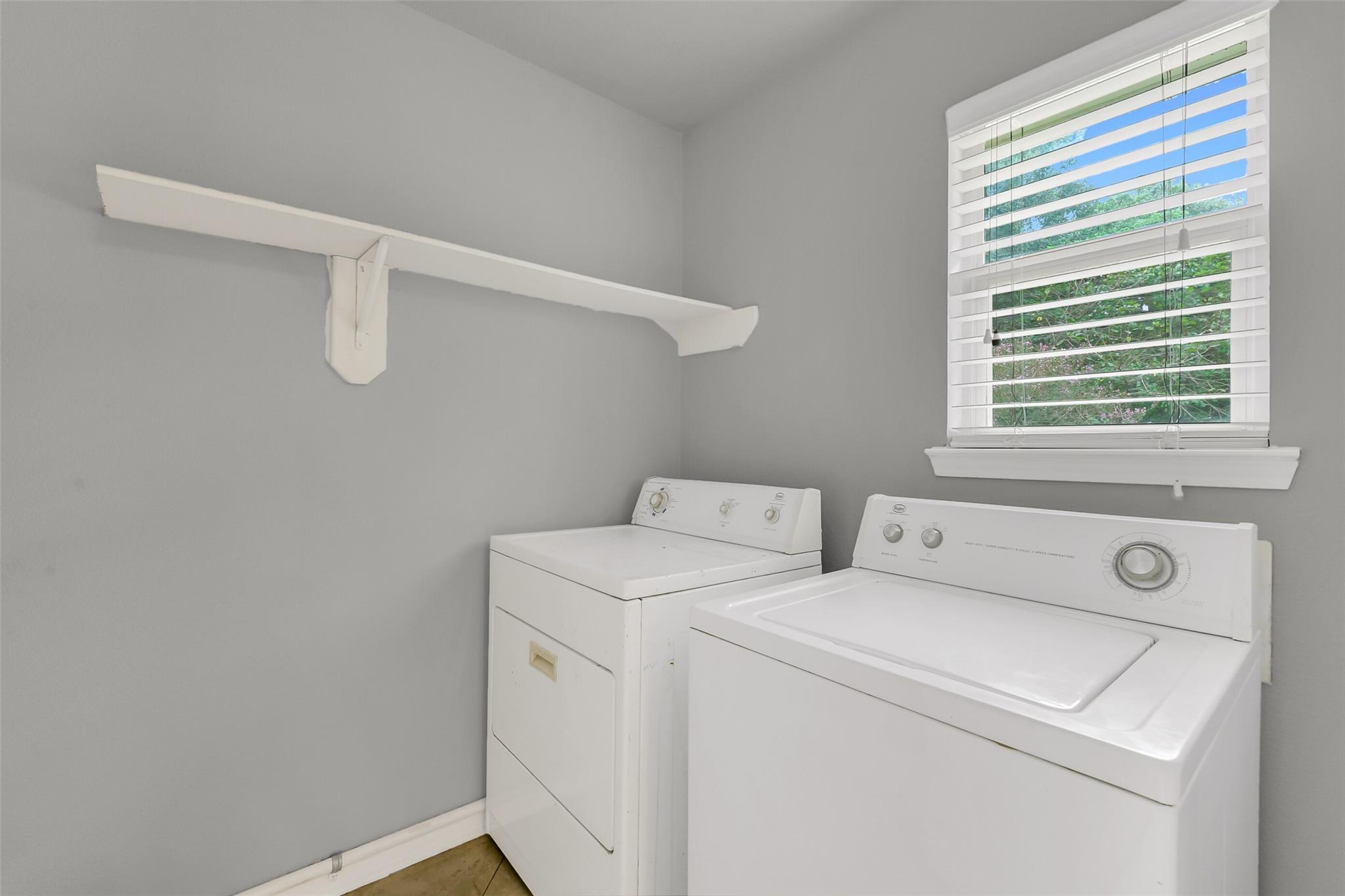 Laundry room with a white washer and dryer beneath a window with blinds.
