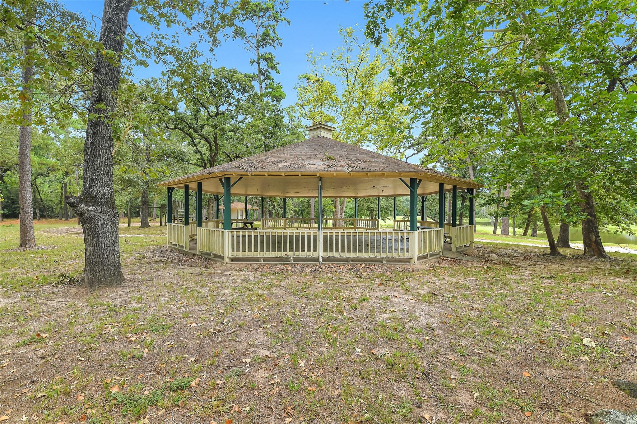 Wooden gazebo with a broad roof and railings beneath mature trees beside a pond.