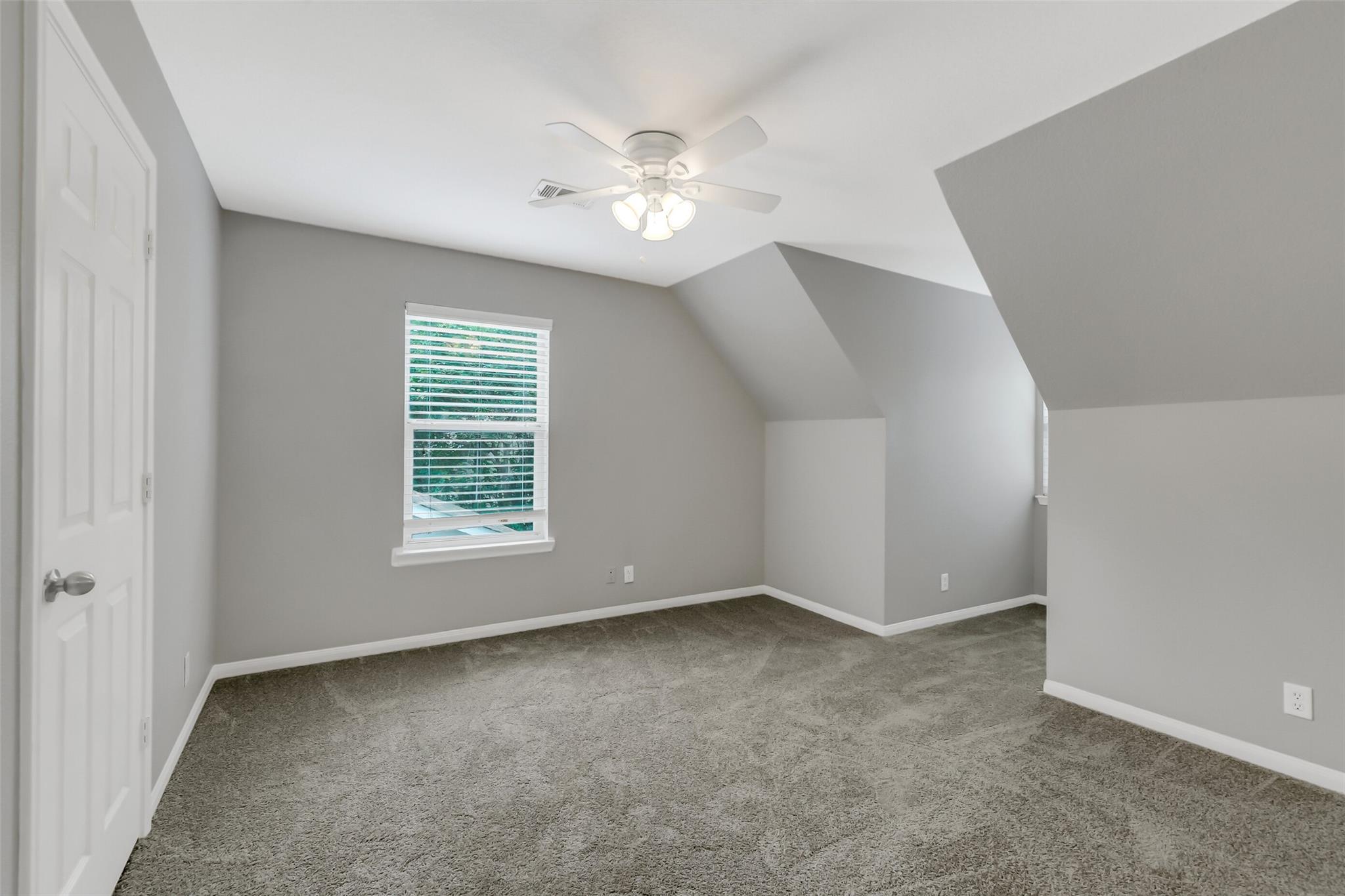 Empty carpeted room with a ceiling fan, sloped ceiling, and window with blinds.