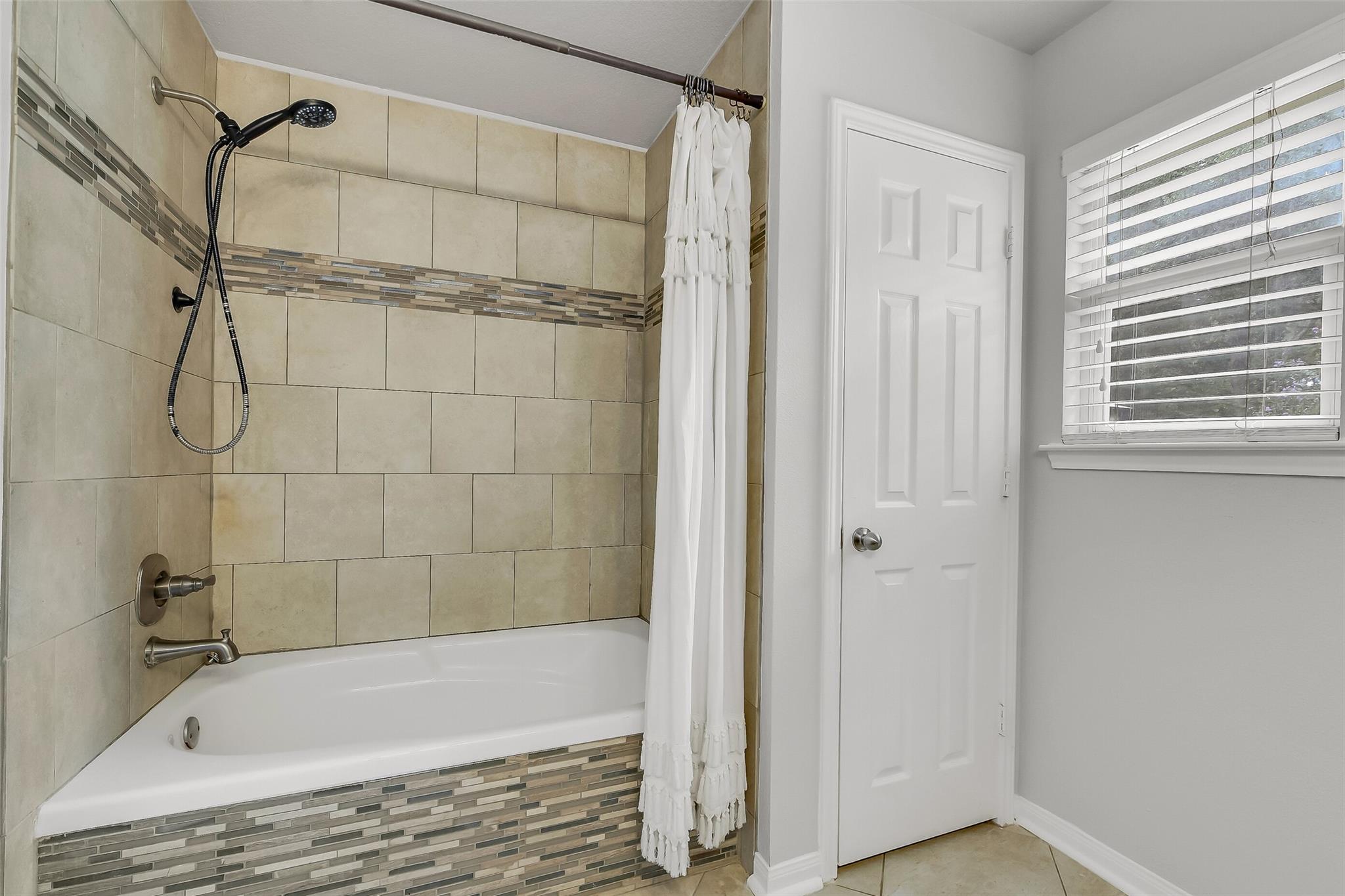 Bathroom with a tiled bathtub and shower, white curtain, and window with blinds.