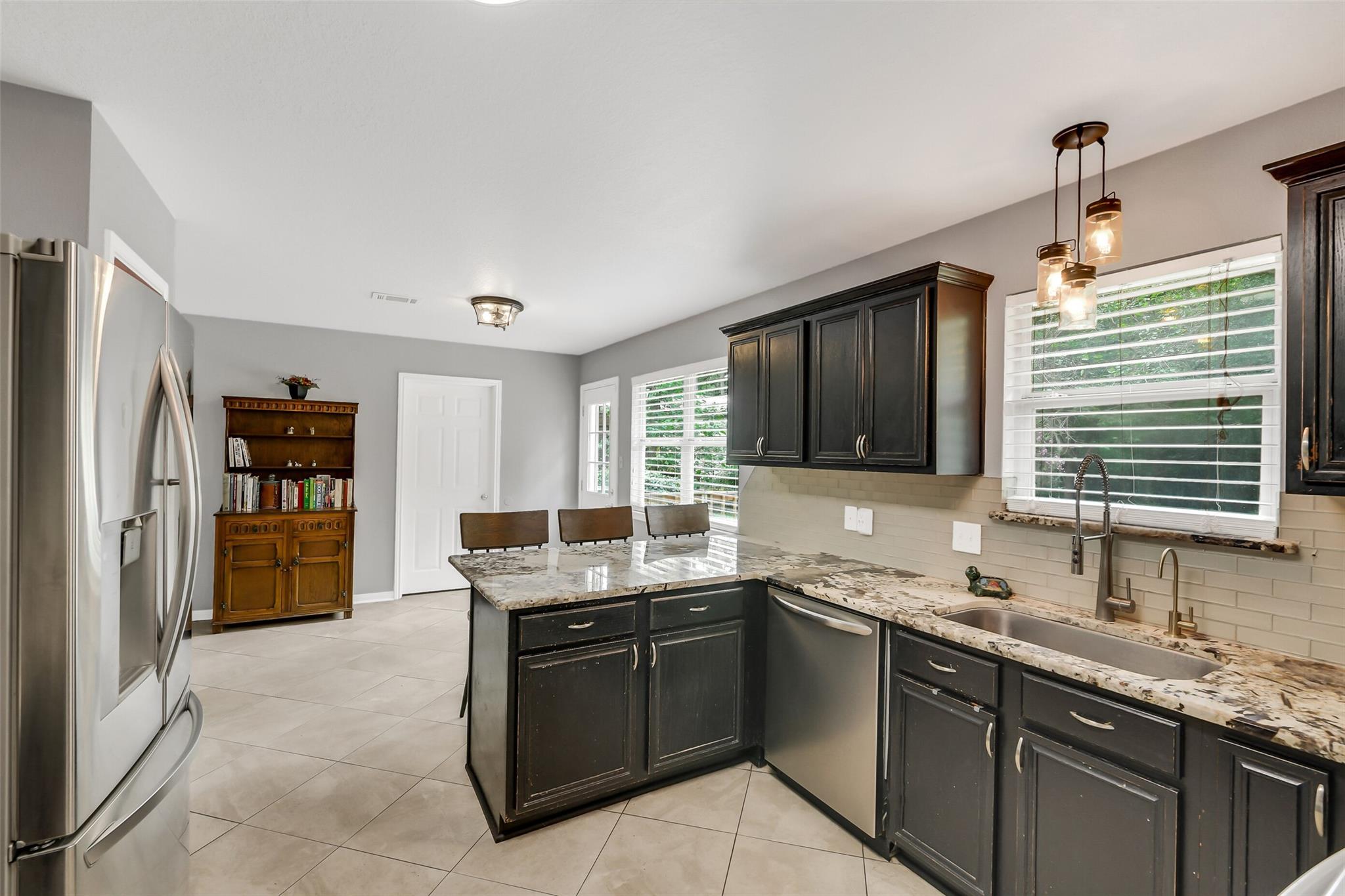 Kitchen with dark cabinets, granite island, stainless steel refrigerator, and windows with white blinds.