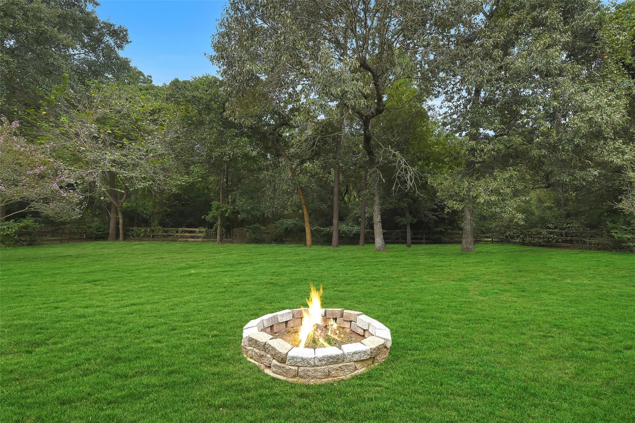 Backyard lawn with a circular stone fire pit, mature trees, and a wooden fence.