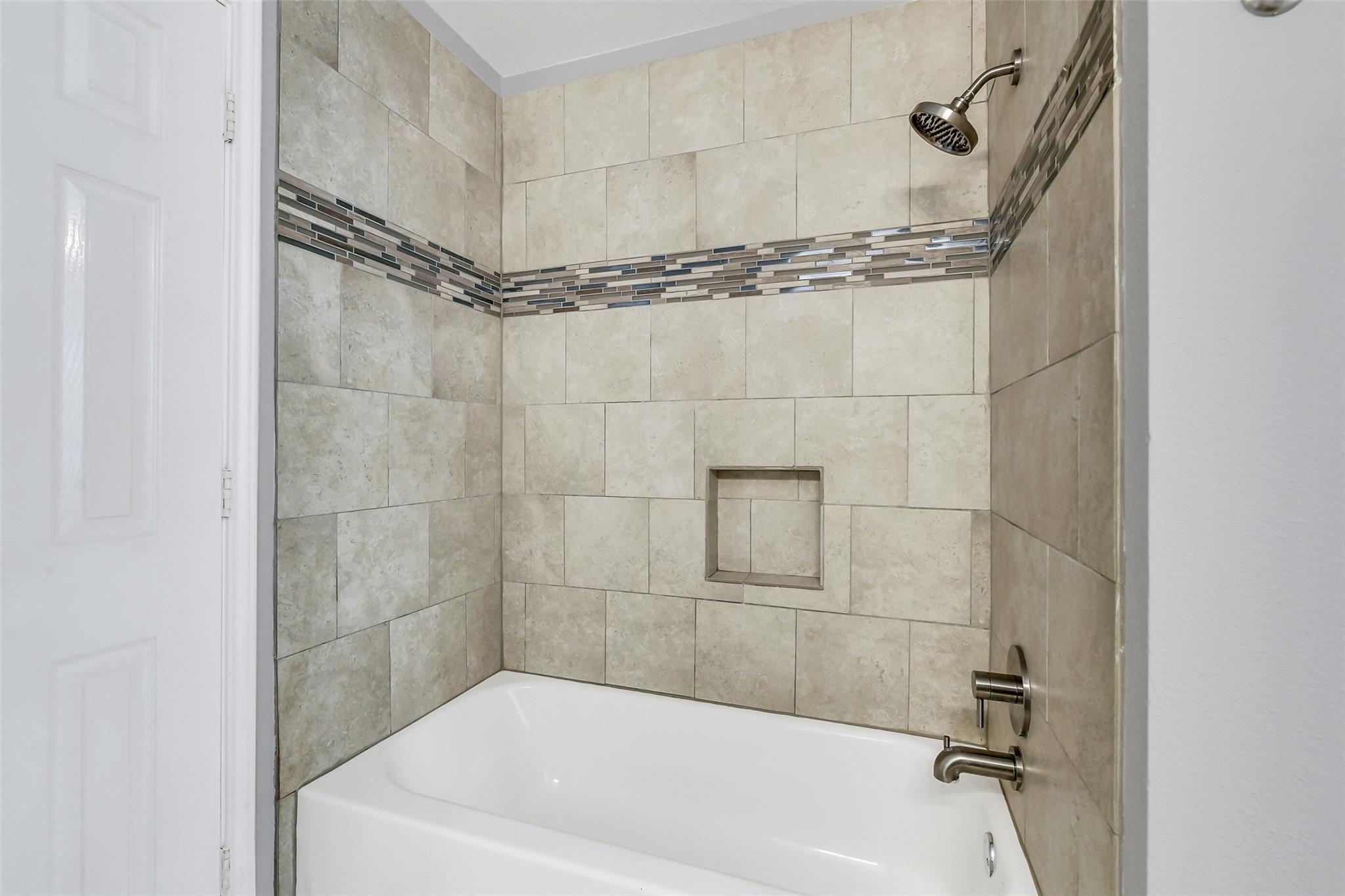 Bathtub with a beige tiled shower surround, mosaic accent band, and recessed niche.