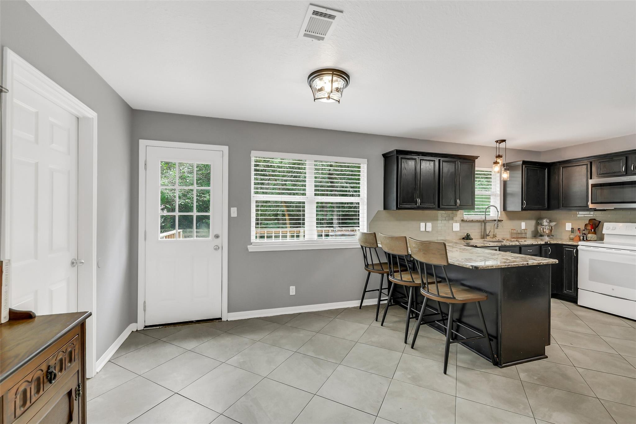 Kitchen with dark cabinets and a stone island with three bar stools, beside a door and windows.