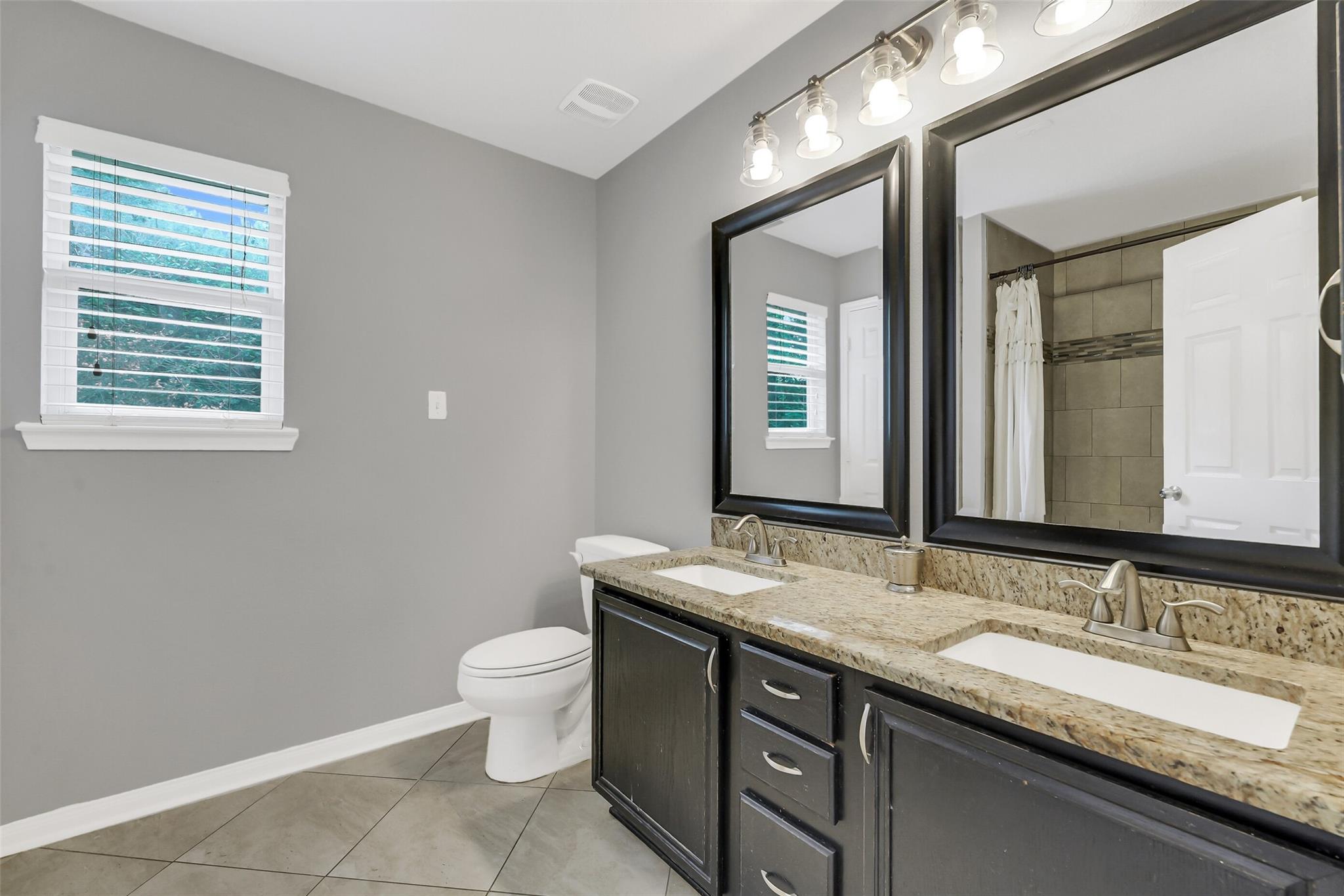 Bathroom with a double-sink granite vanity, dark cabinetry, and a tiled shower.