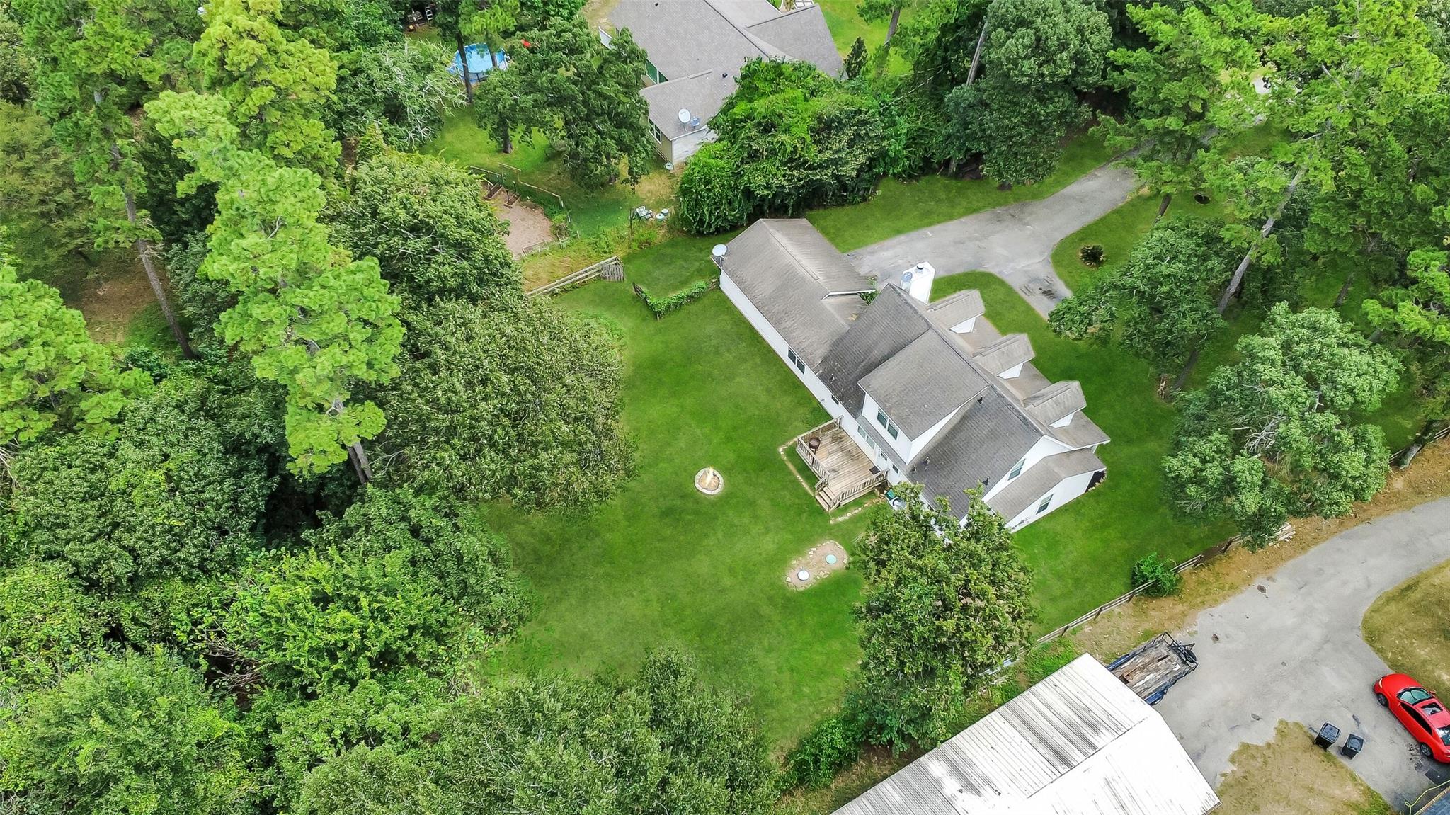 Aerial view of a white house surrounded by mature trees, with a broad lawn, wooden deck, and fire pit.
